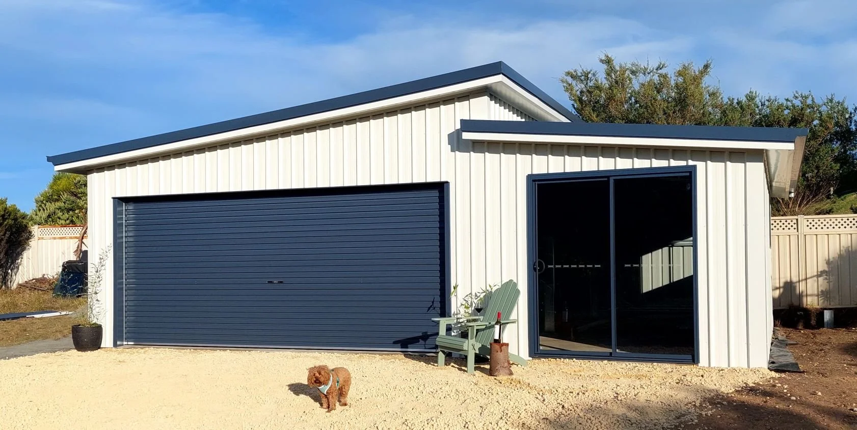 A modern small garage with a closed dark blue roll-up door and a sliding glass door on the right. There is a green Adirondack chair with a small table beside it, a potted plant, and a small dog standing on a gravel surface in the foreground. The back