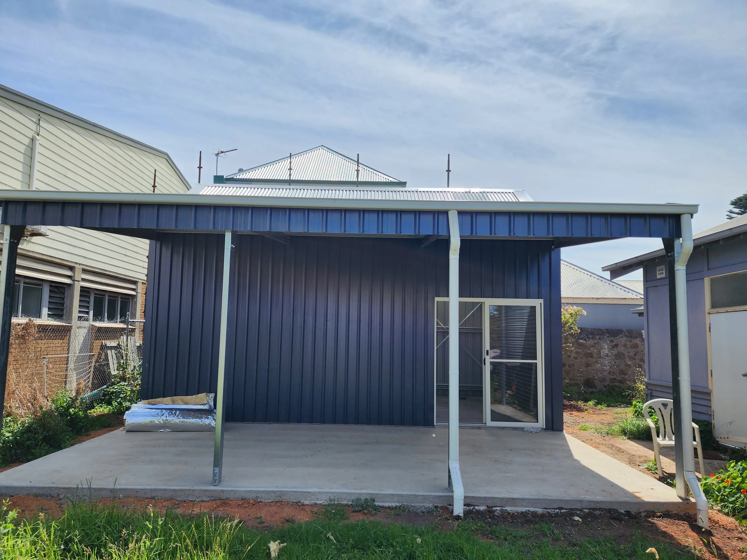 Backyard view of a small shed with a blue metal exterior, sliding glass door, and partial covered patio, built by M&C Sheds