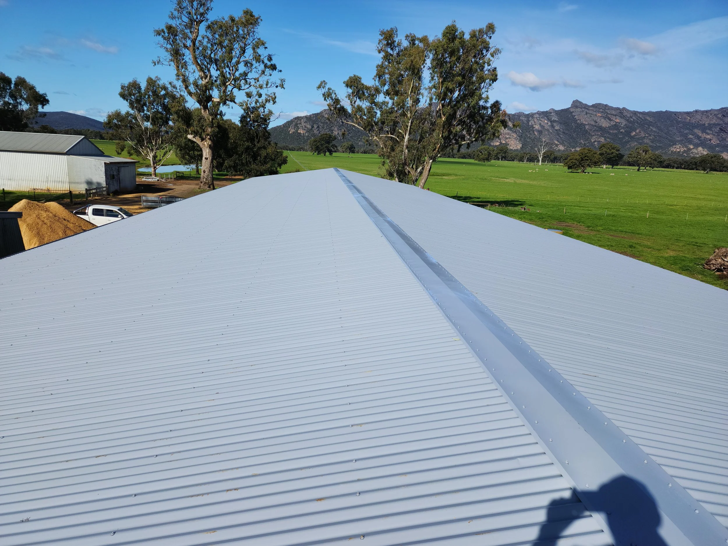 Metal roof with a ridge vent, surrounded by green fields, trees, a barn, a parked truck, and mountains in the background under a blue sky with scattered clouds.