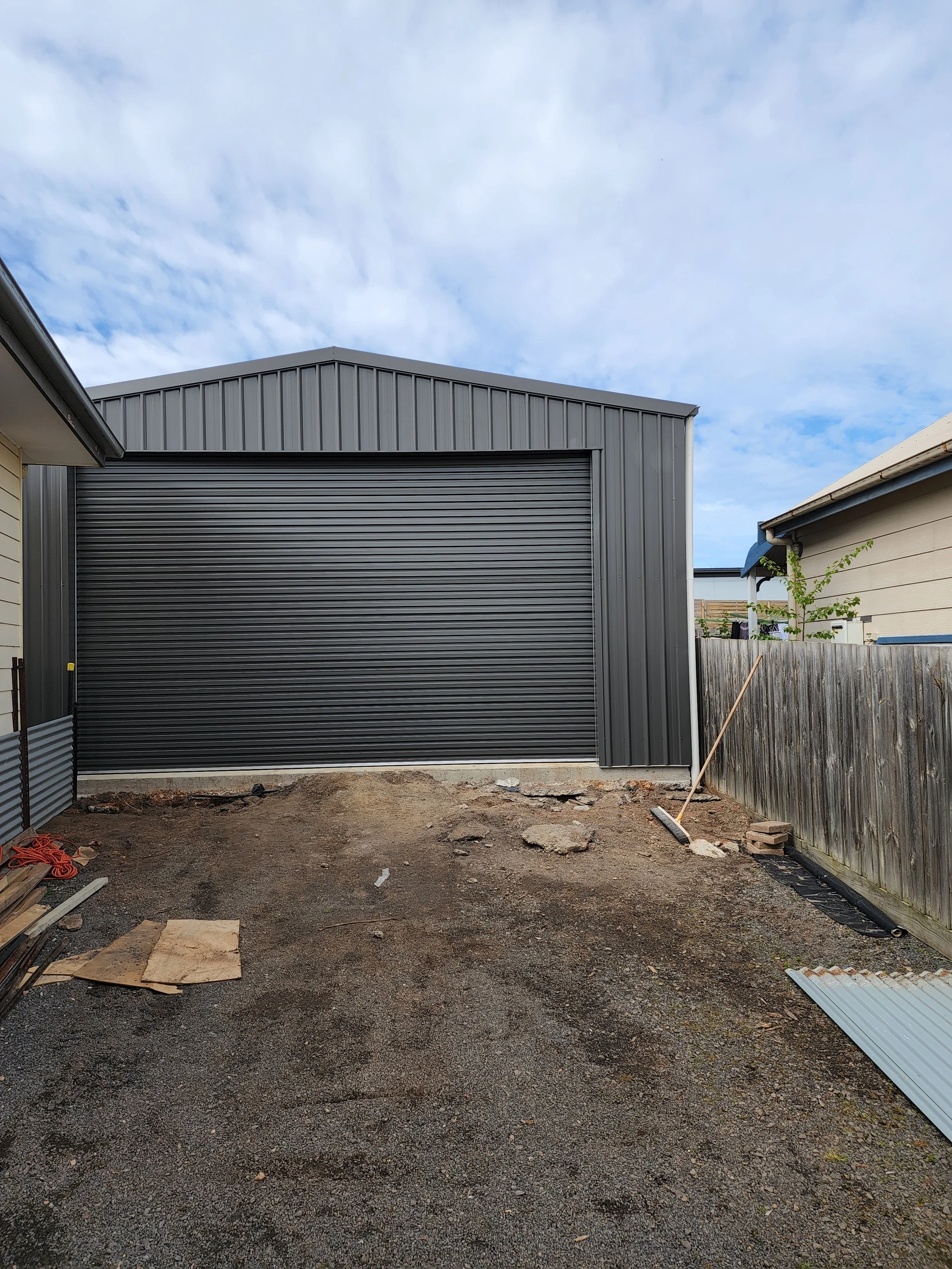 Backyard with a new grey metal shed, dirt ground, wooden fence, and a cloudy sky.