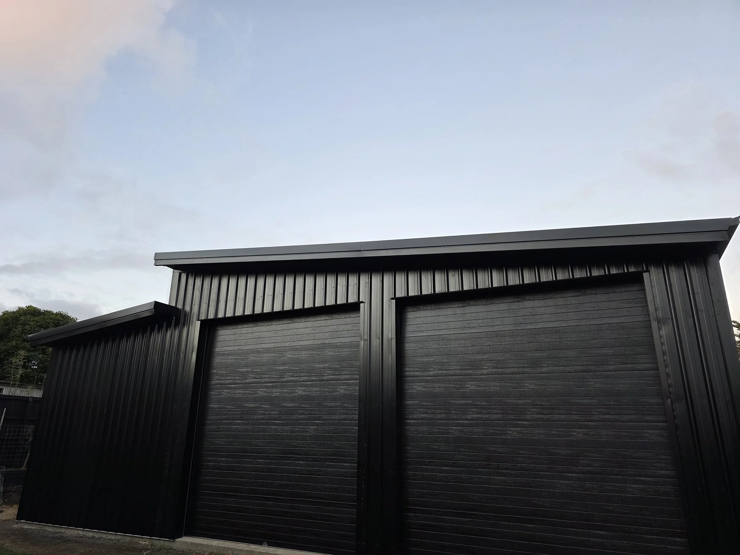 A black metal storage shed or garage with roller doors under a cloudy sky.