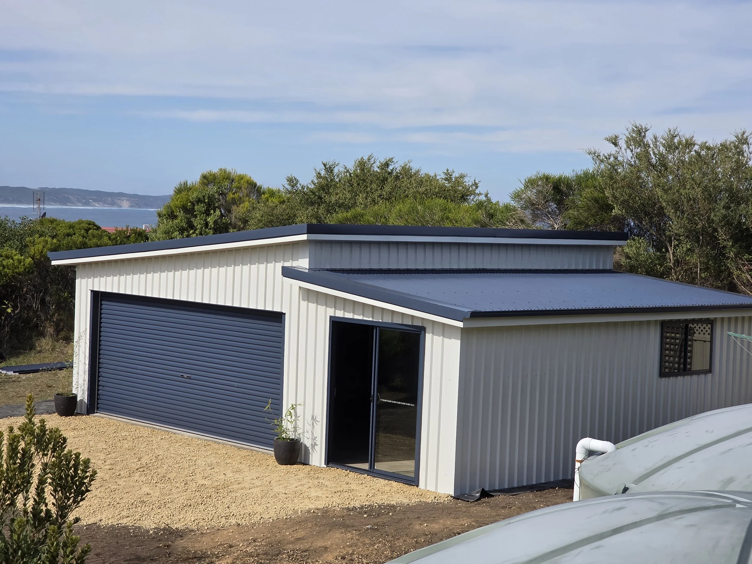 Metal workshop with a black roller door, sliding glass door, and small window, set in a gravel yard with potted plants, overlooking a landscape with trees, water, and hills under a partly cloudy sky.