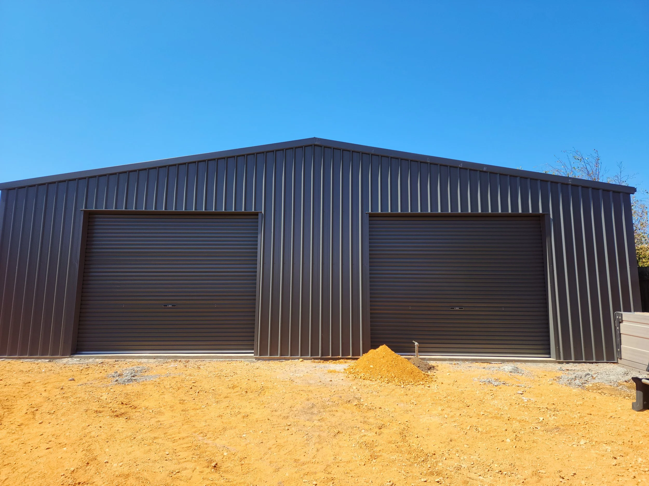 A black metal building with two closed roll-up garage doors, situated on a dirt lot under a clear blue sky.