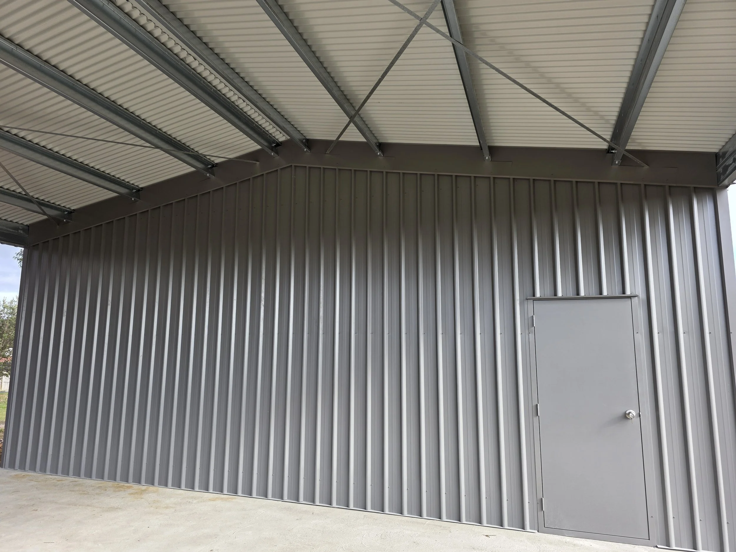 Exterior of a metal shed with a corrugated metal wall and a gray door built by M&C Sheds