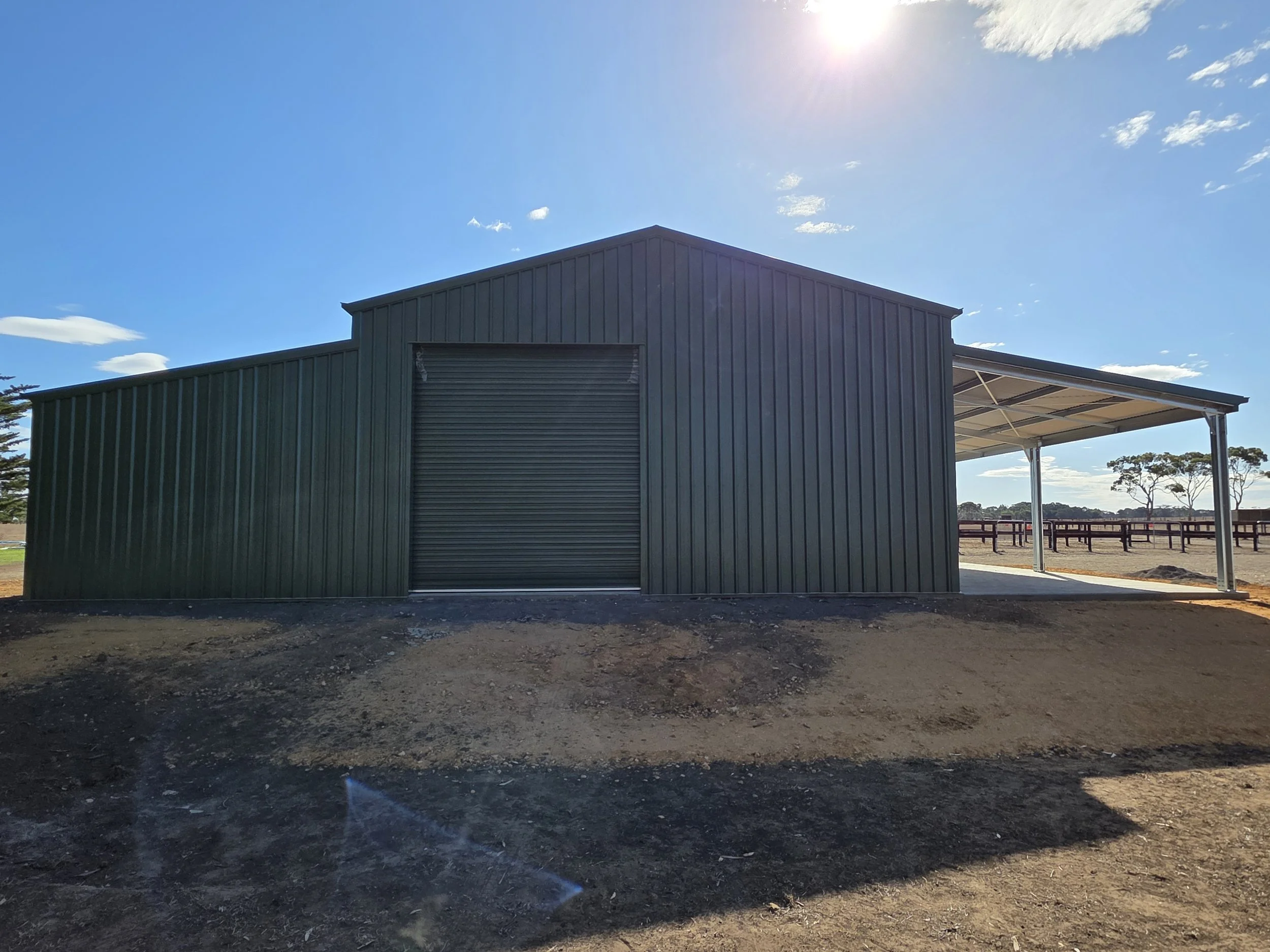 A green metal shed outdoors with a closed roll-up door and open side area under a sloped roof, built by M&C Sheds
