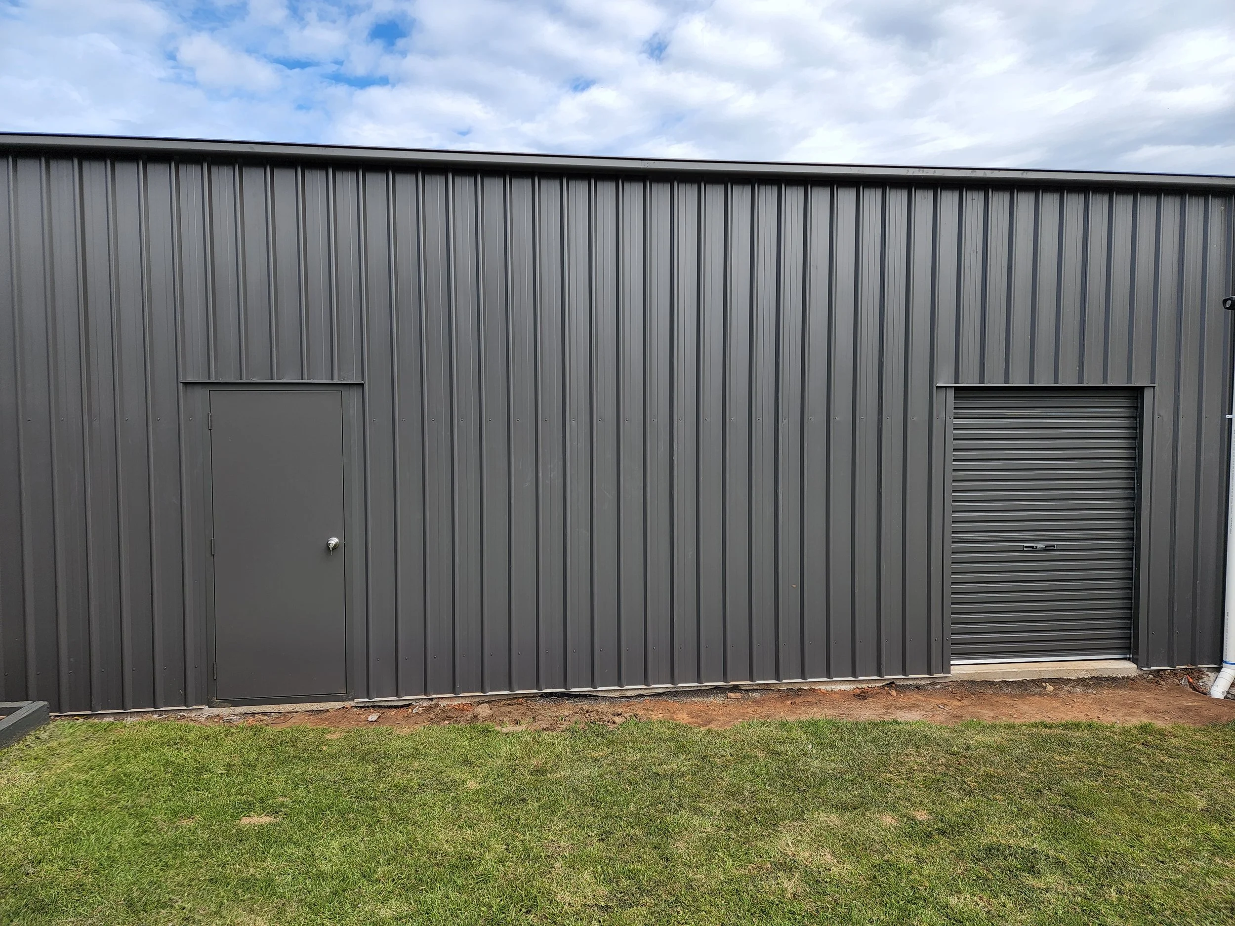 Exterior of a gray metal building with a door on the left and a roll-up garage door on the right, with grass and dirt in front, and cloudy sky above.