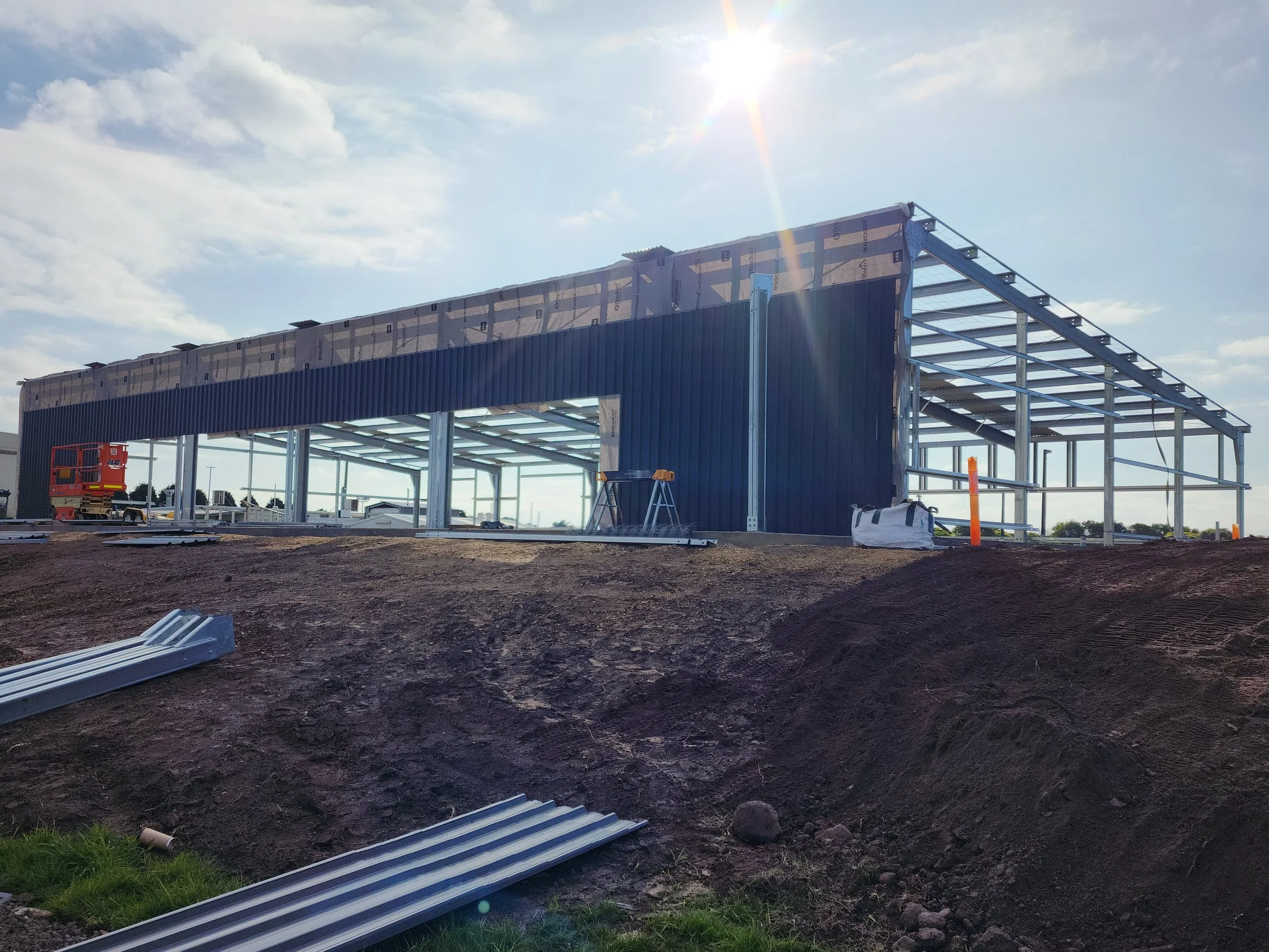 Steel framework of a building under construction with a partially installed exterior wall, situated on a dirt lot under a sunny sky.