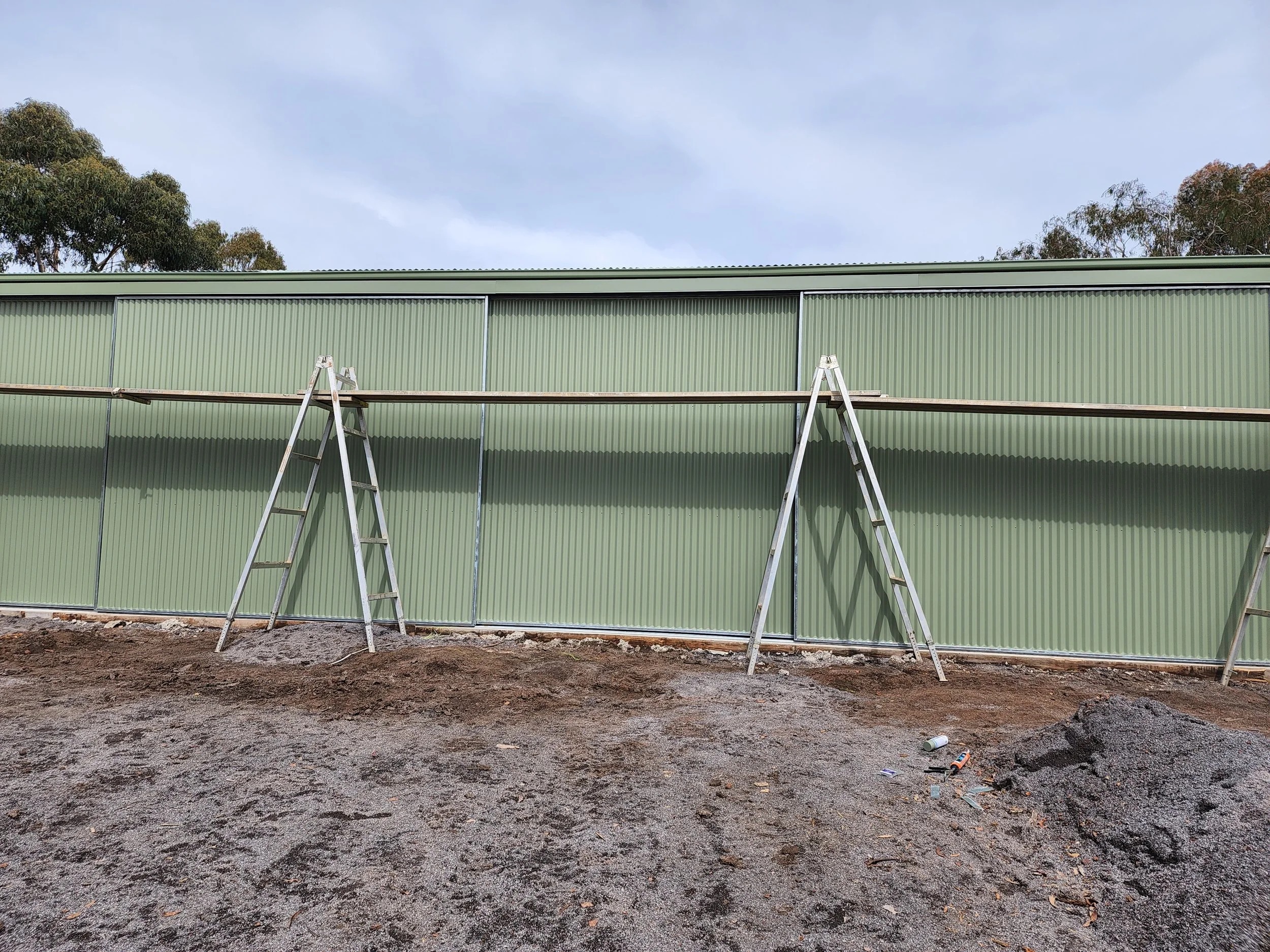 Construction site with two metal ladders leaning against a green corrugated metal wall, dirt ground with some debris and a pile of gravel, trees in the background under a cloudy sky.