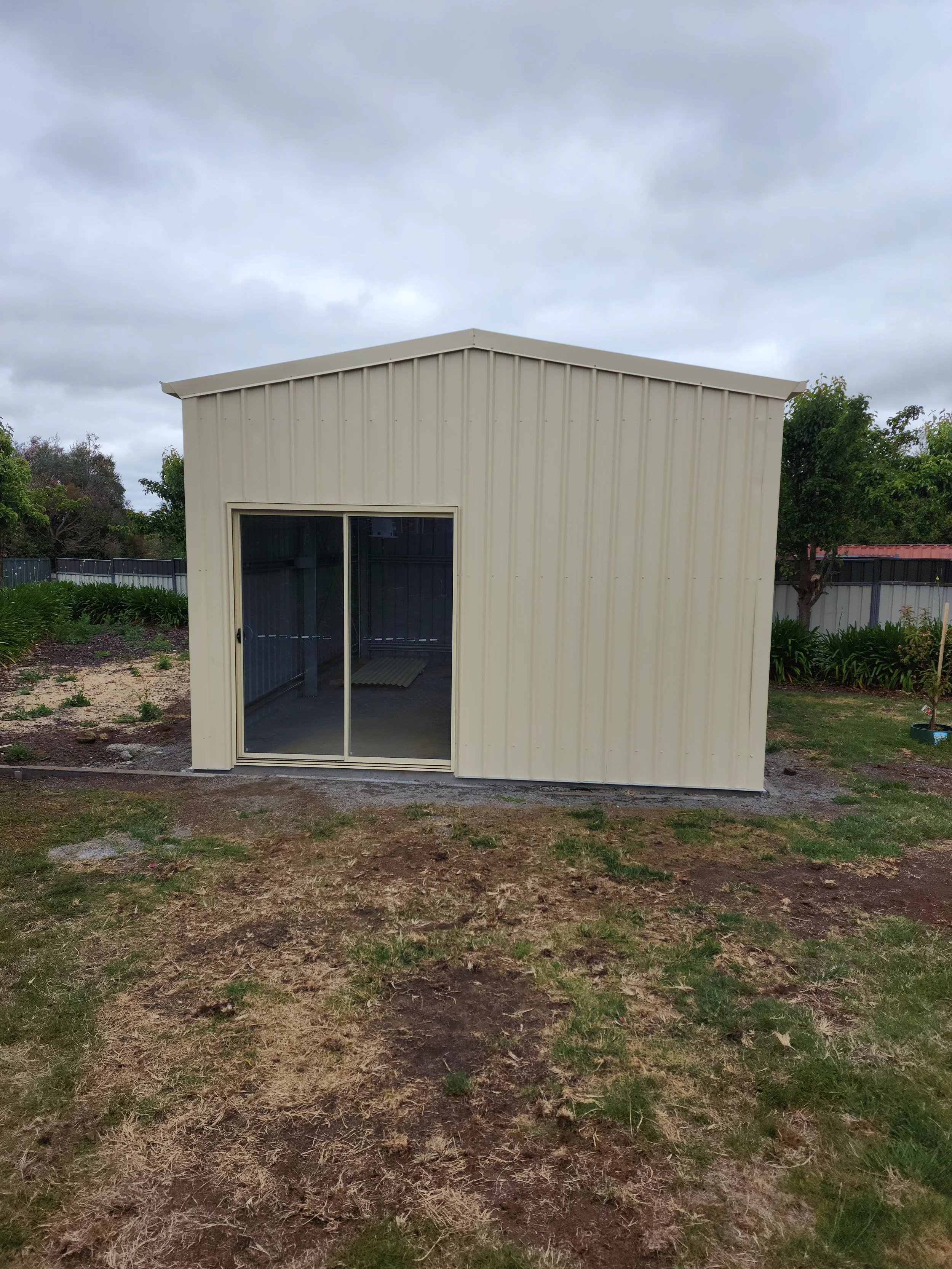Small beige metal shed with sliding glass door in a backyard built by M&C Sheds