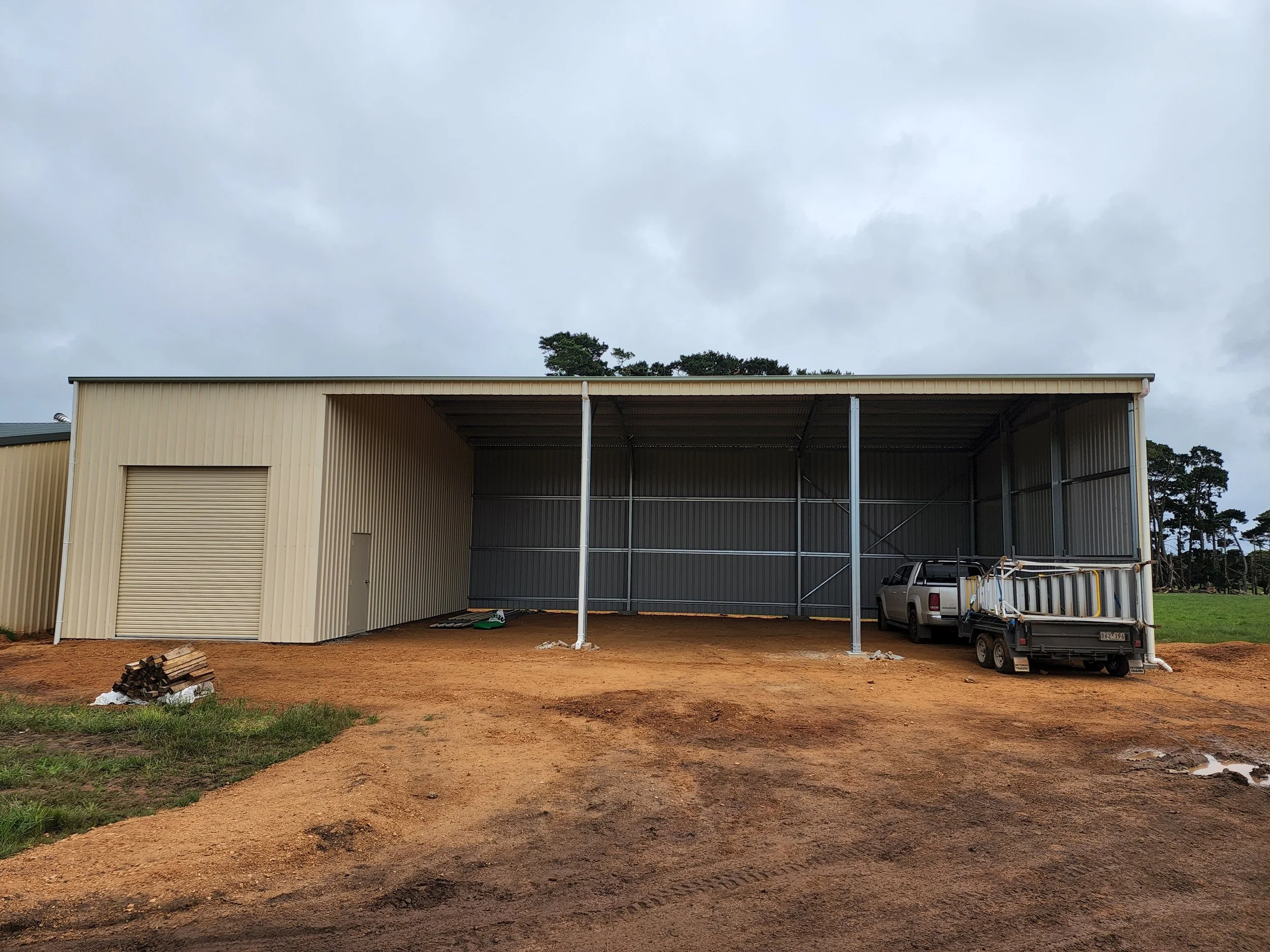 A metal storage shed with large sliding door, small side door, open area inside, parked pickup truck with trailer, built by M&C Sheds