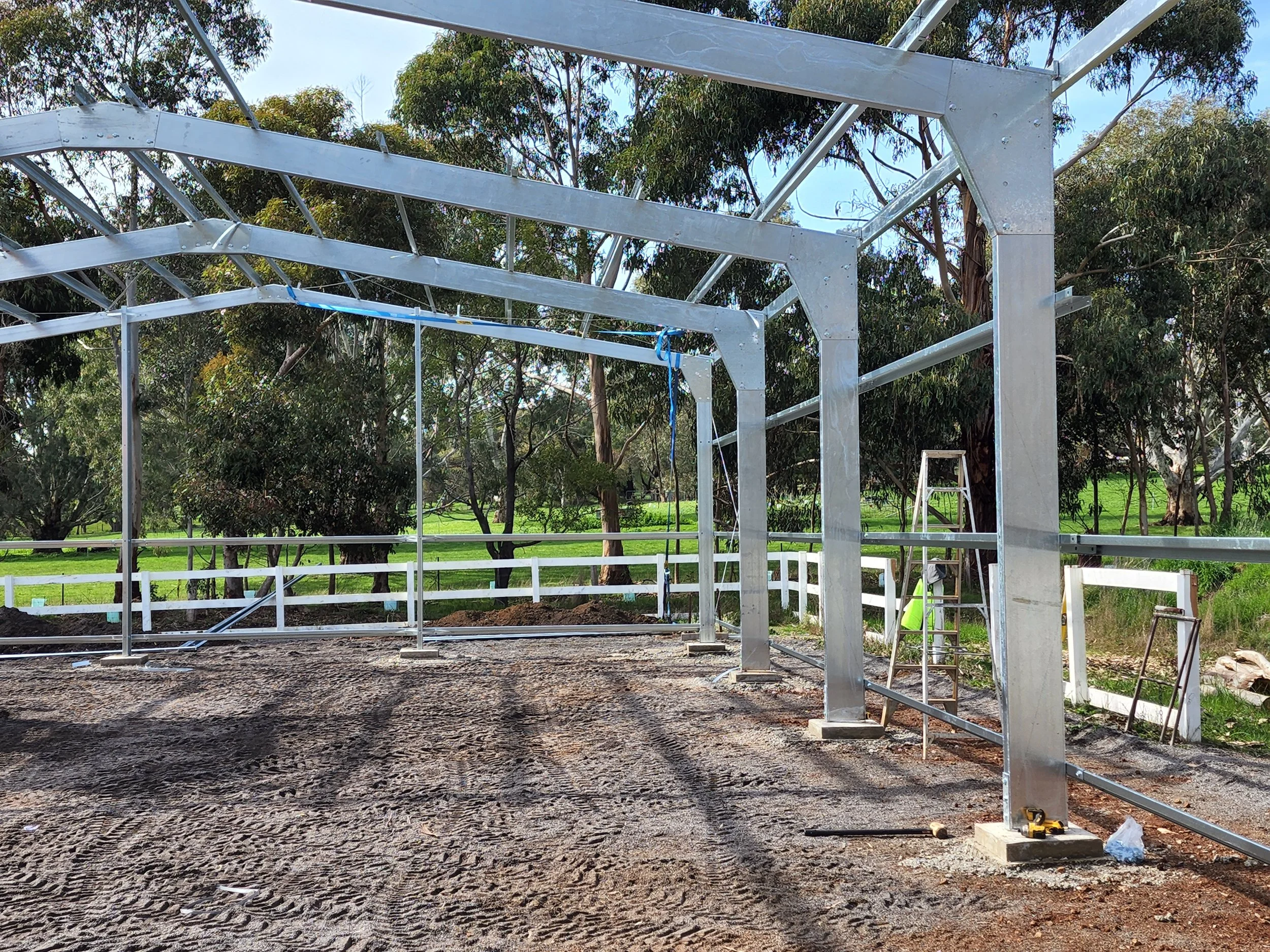 Construction of a metal frame structure, possibly for a building, with a ladder and construction tools on the ground. The structure is outdoors surrounded by trees and a white fence.
