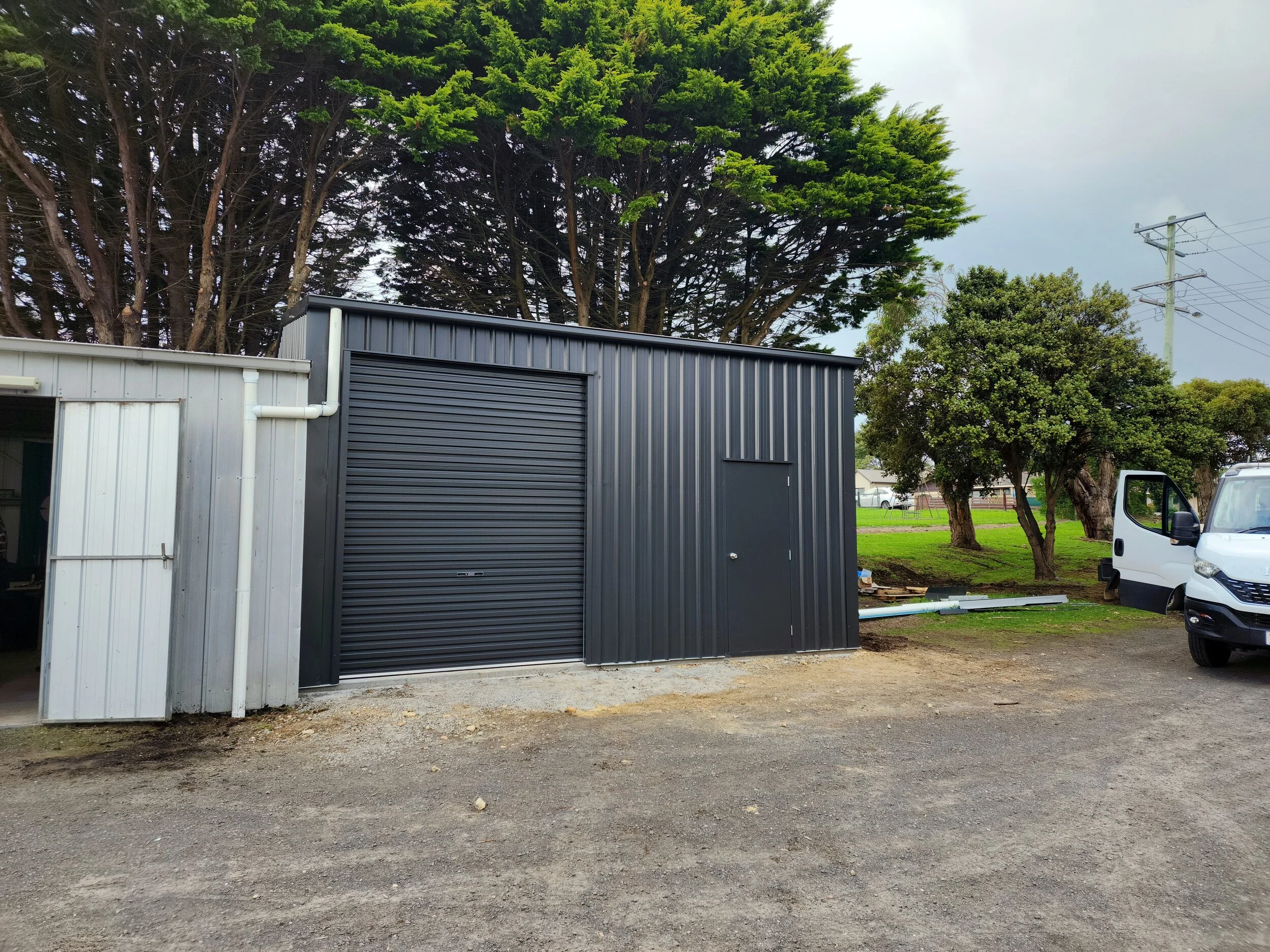 A small black metal storage shed with a roll-up door and a side door, built by M&C Sheds