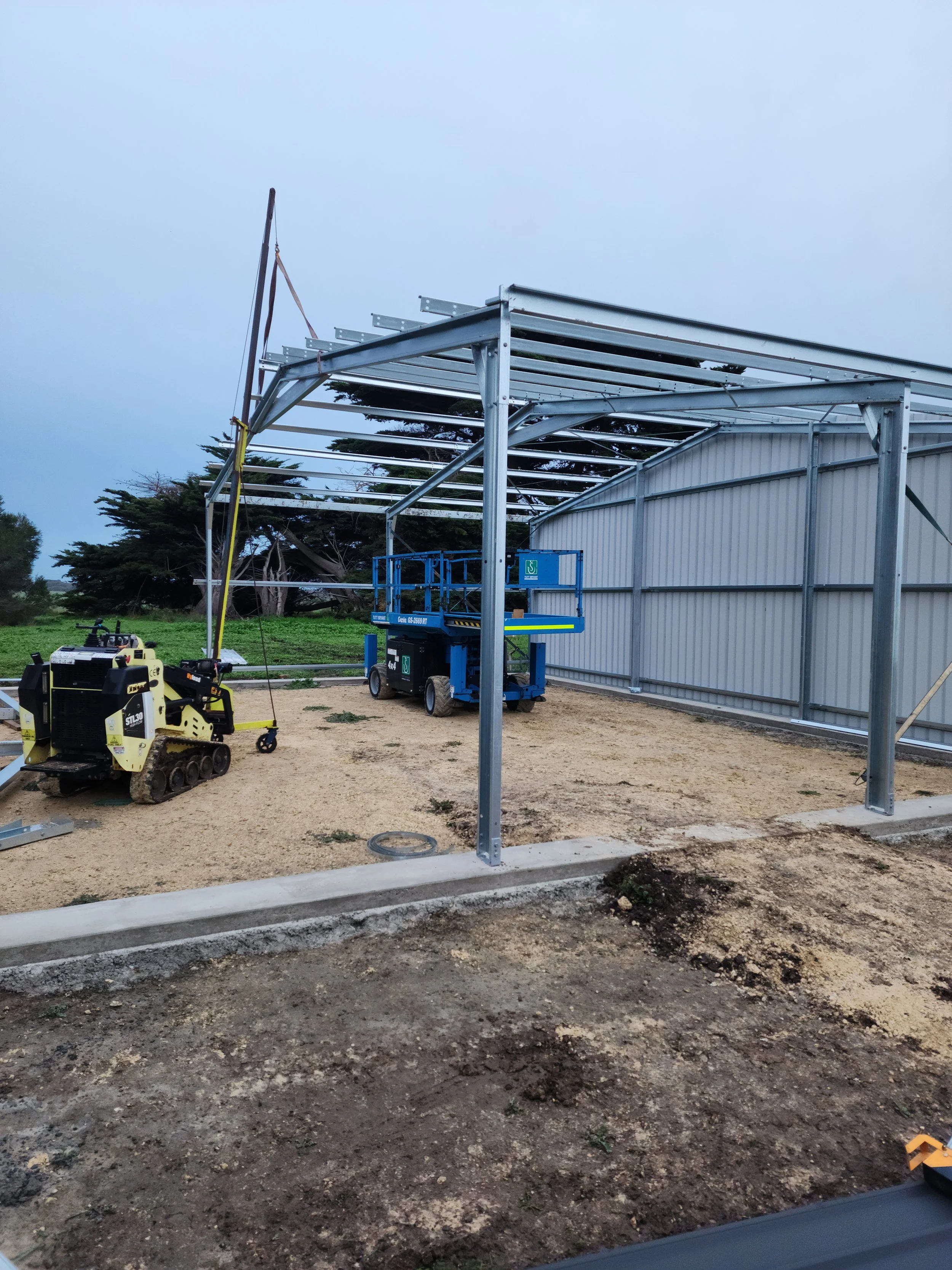 Construction site with metal framework, construction equipment, and a partially built structure outdoor shed built by M&C Sheds