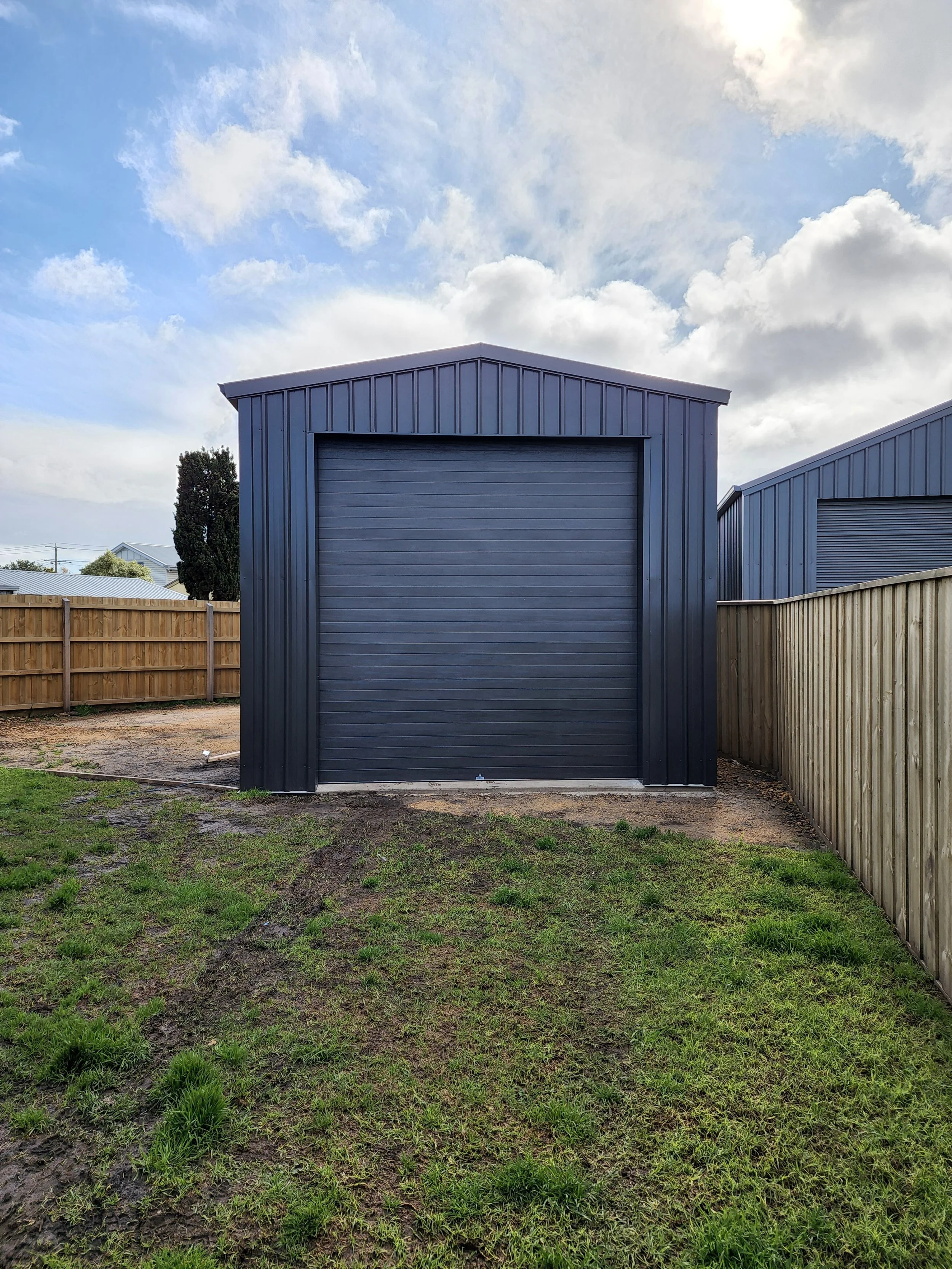 A black metal shed with a roll-up door in a backyard, surrounded by a wooden fence, with partly cloudy sky overhead.