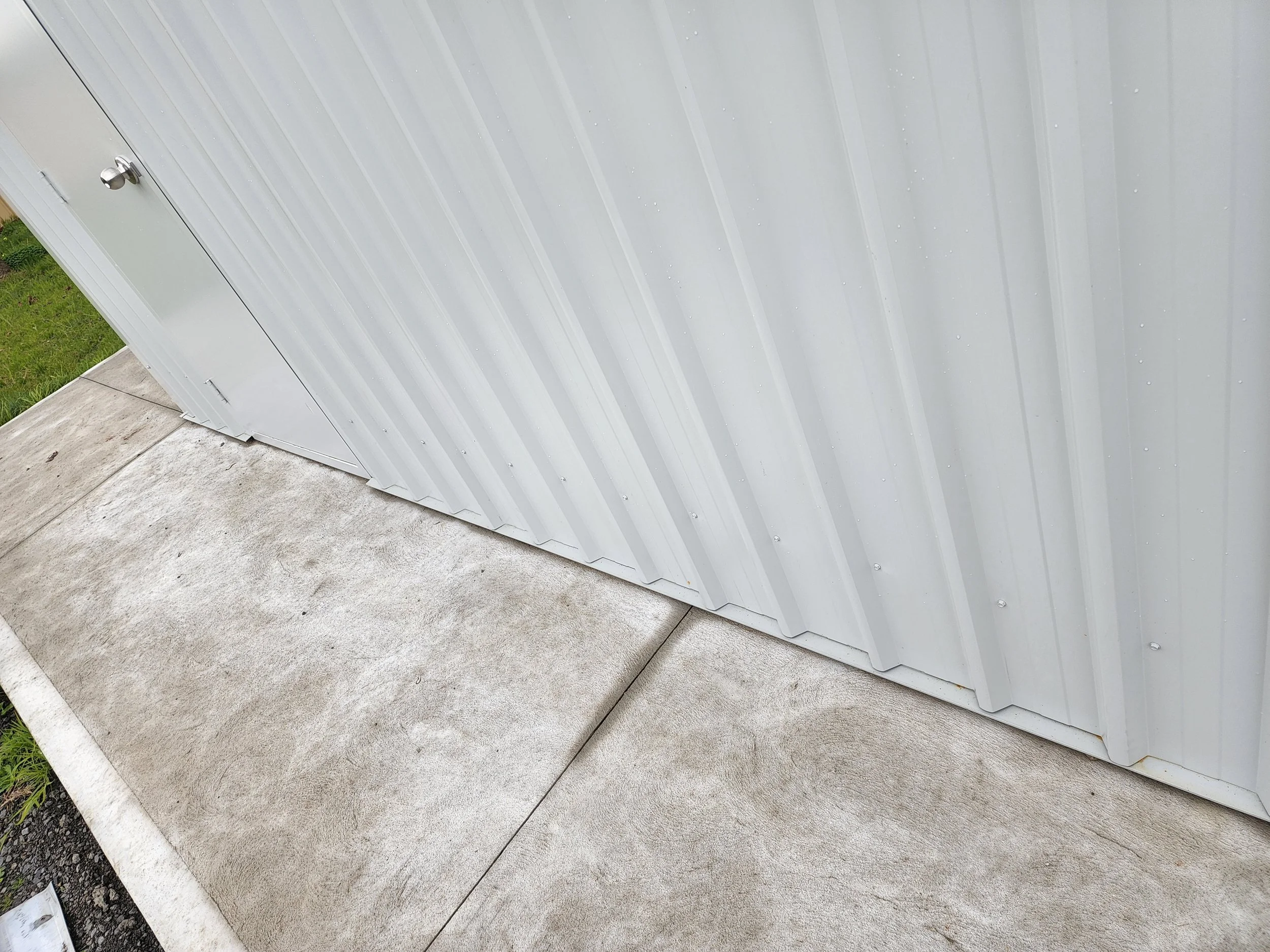 Close-up of a light gray metal building exterior with vertical ridges, situated on a concrete slab with grass visible nearby.