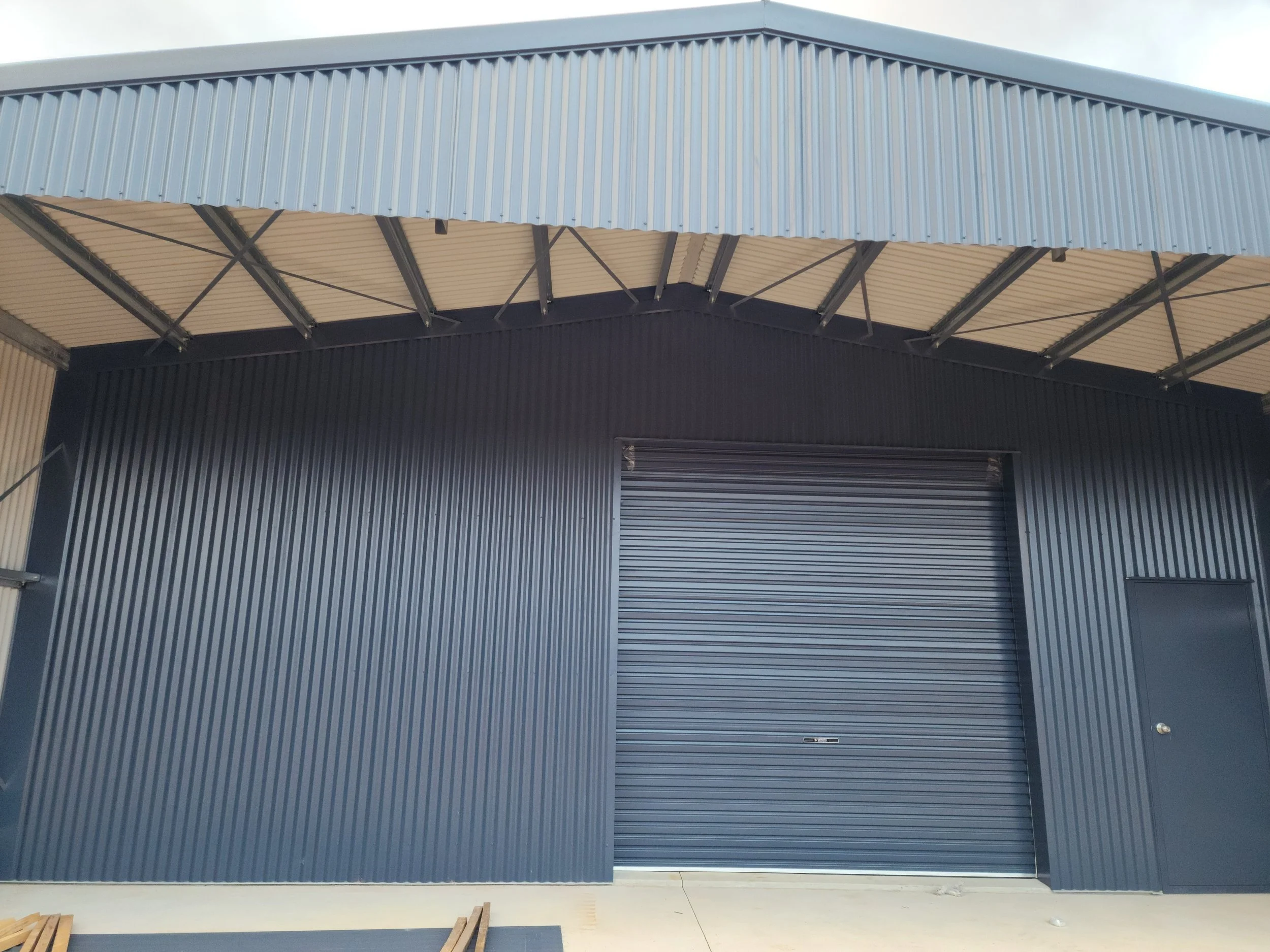 Exterior view of a modern industrial warehouse shed with a large blue roll-up door and side door, constructed from corrugated metal panels.