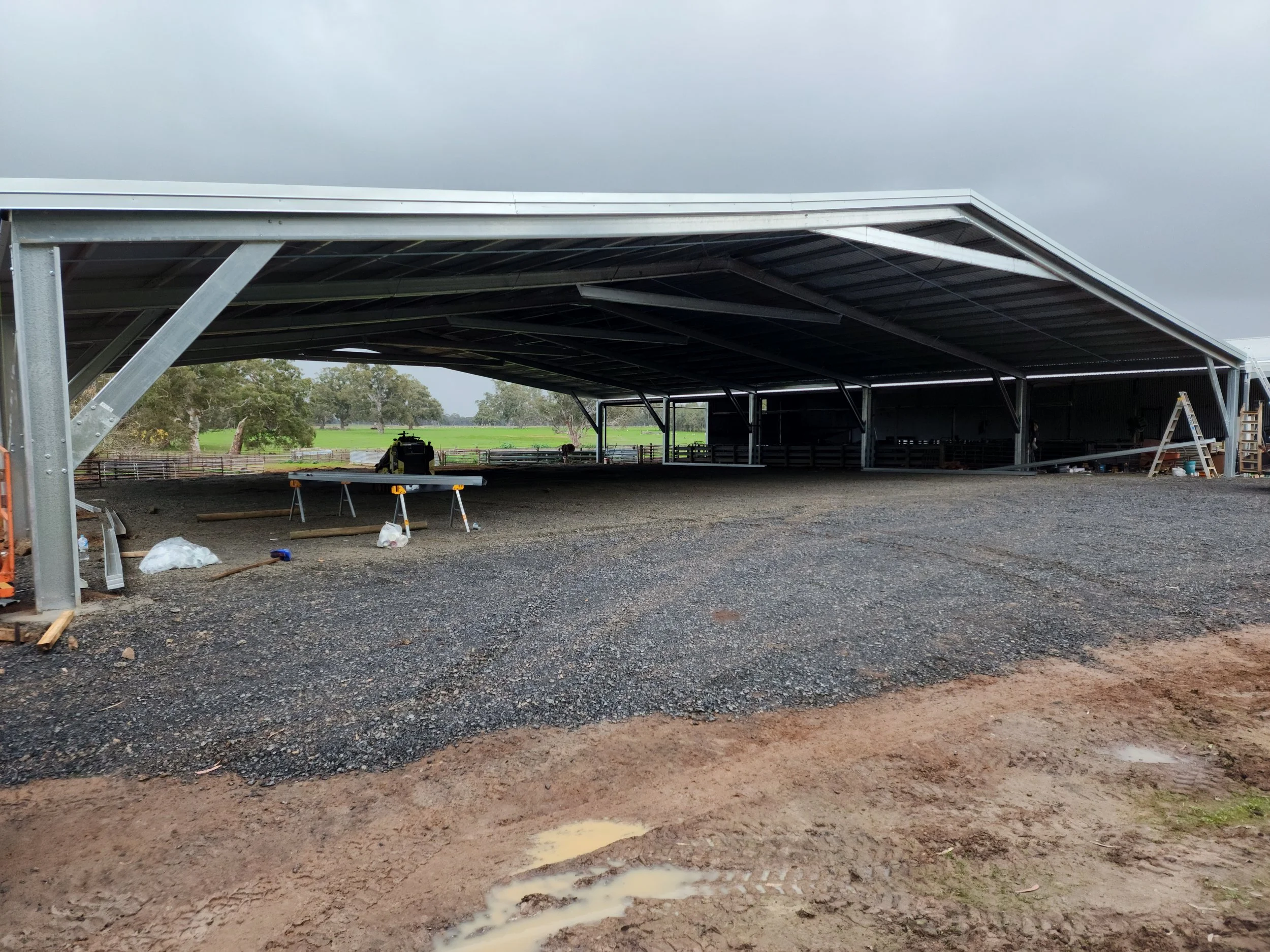 Construction site of a large metal structure or barn with a curved roof, partially built, on a rural property with fields and trees in the background, cloudy sky overhead.