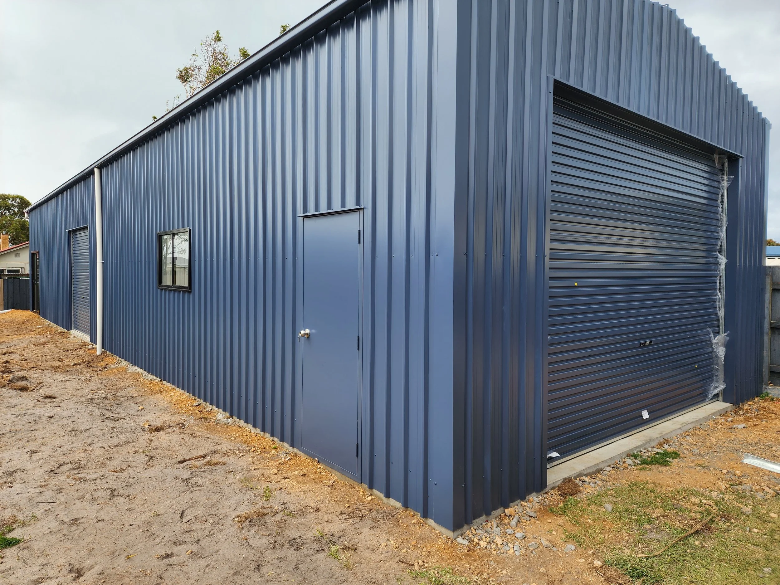 Blue metal storage shed with a side door, small window, and partially closed rolling shutter door, built by M&C Sheds