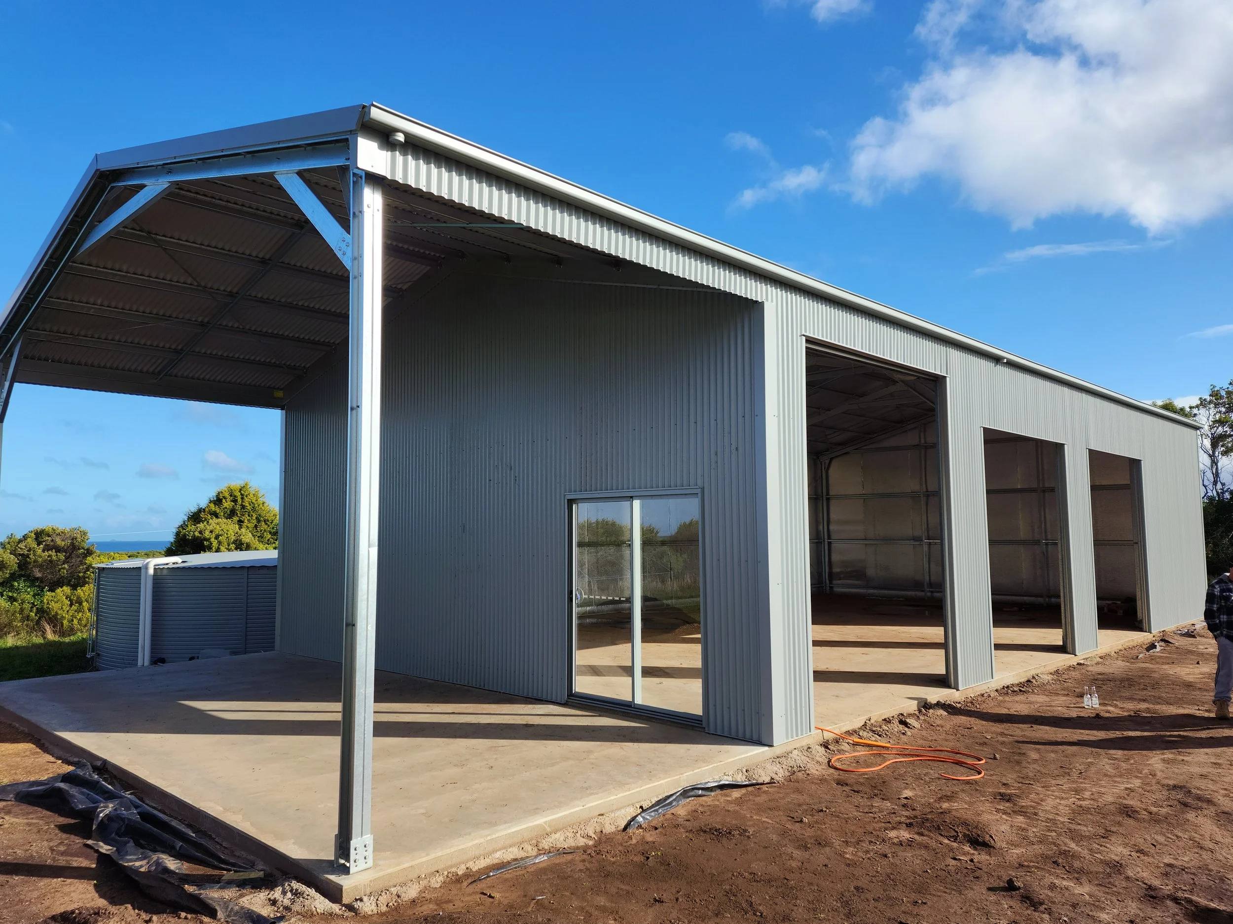 A partly constructed metal building with a concrete pad, open sections, and sliding glass door, set outdoors on dirt with some construction materials around, under a blue sky with a few clouds.