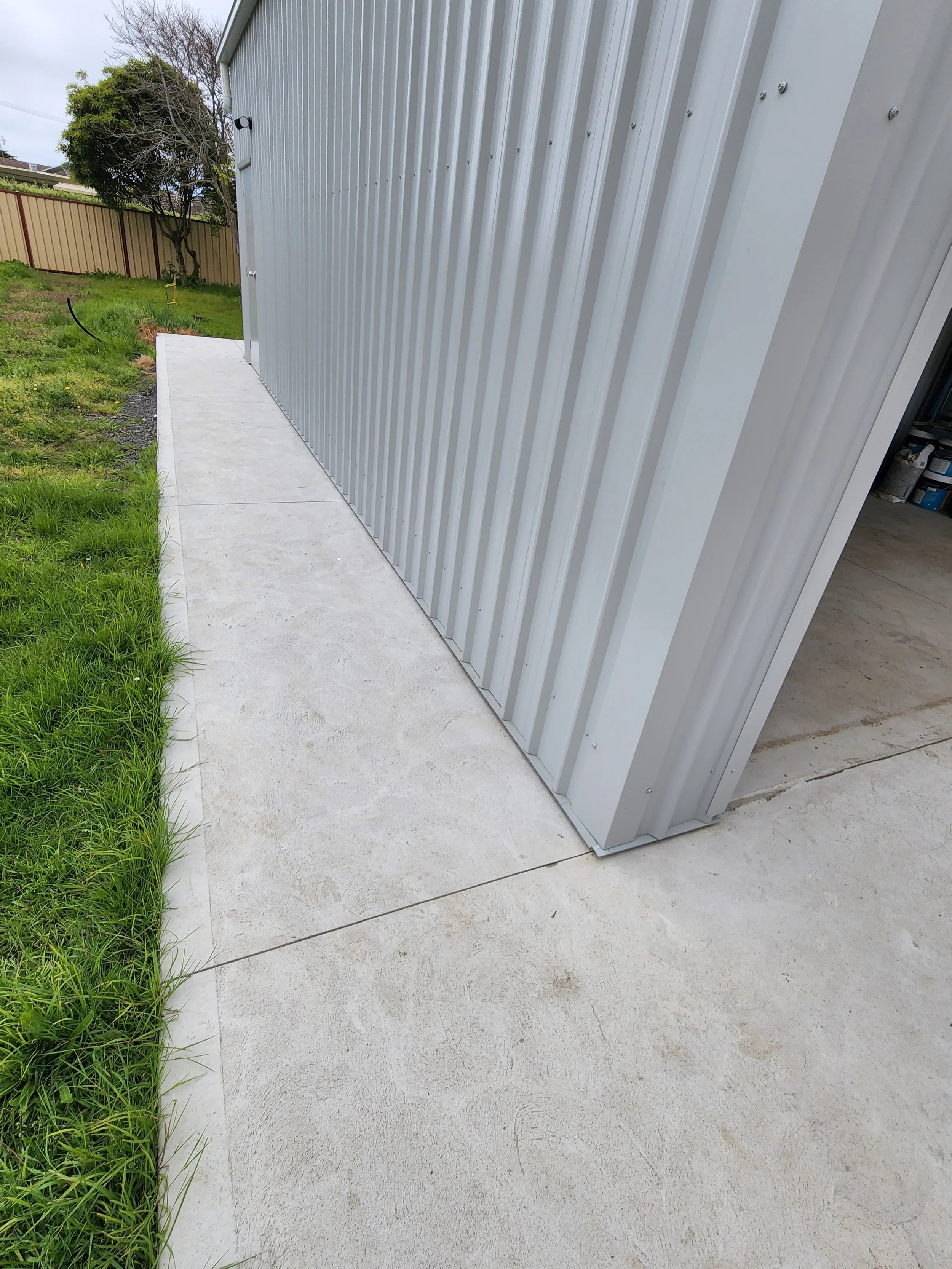 Concrete sidewalk next to a metal building with vertical siding, grass on the left, and a tree in the background.