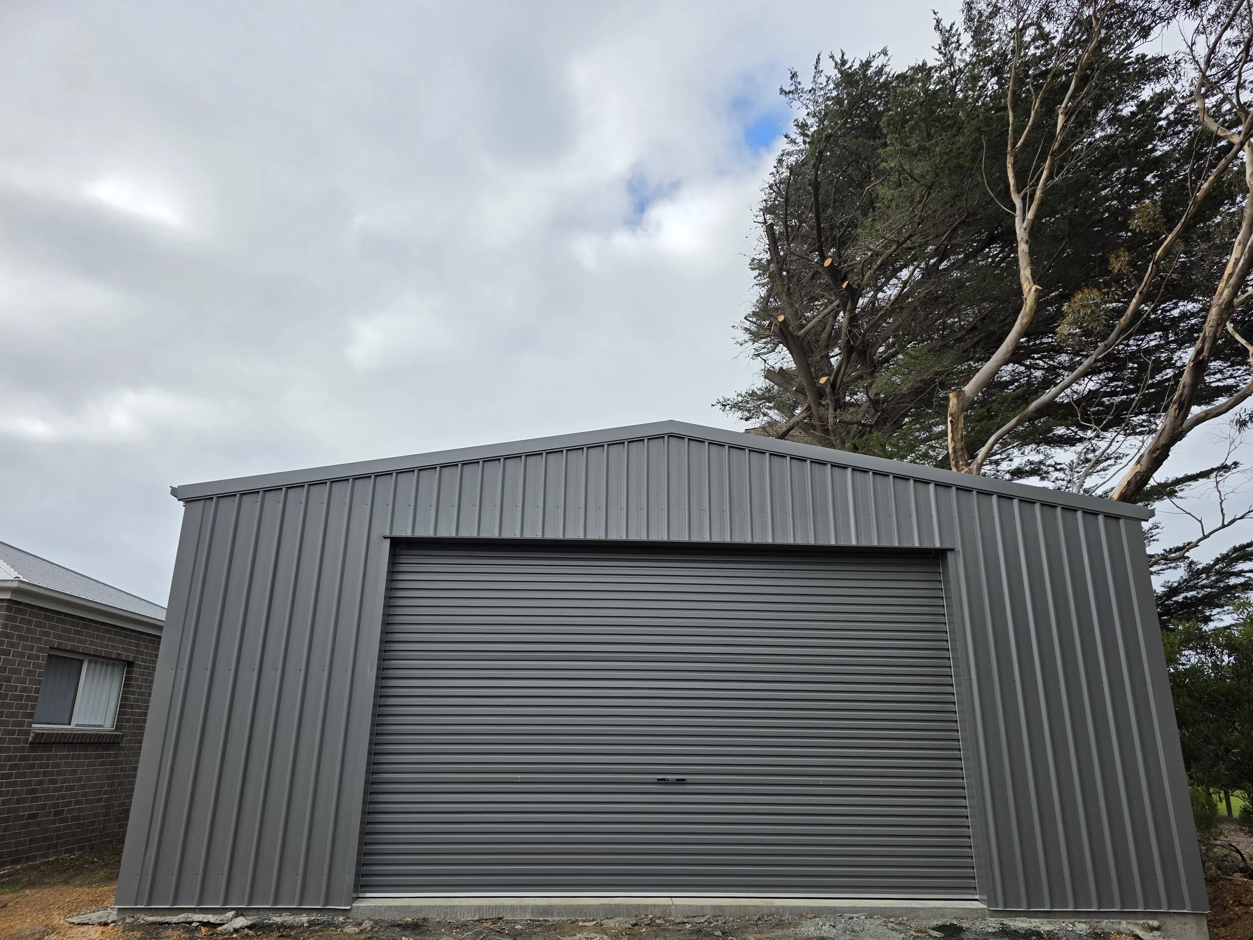 A gray metal storage shed with a rolling door, situated outdoors near trees and a neighboring brick house, under a cloudy sky.