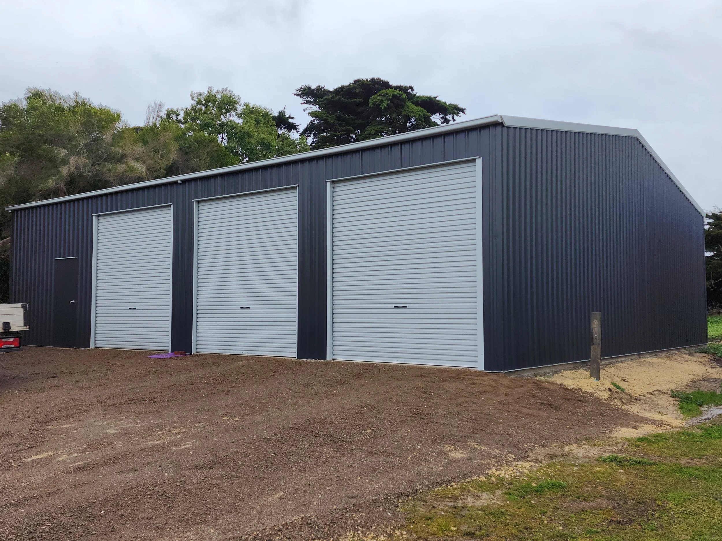 A large, gray metal storage building with three garage doors and one smaller door on an unpaved lot with grass on the edges, surrounded by trees.