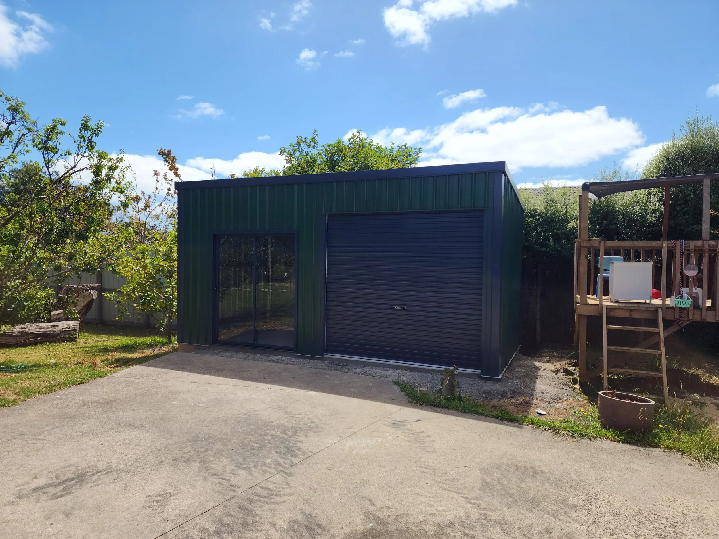 A metal green storage shed with a sliding glass door and a roll-up garage door in a backyard built by M&C Sheds