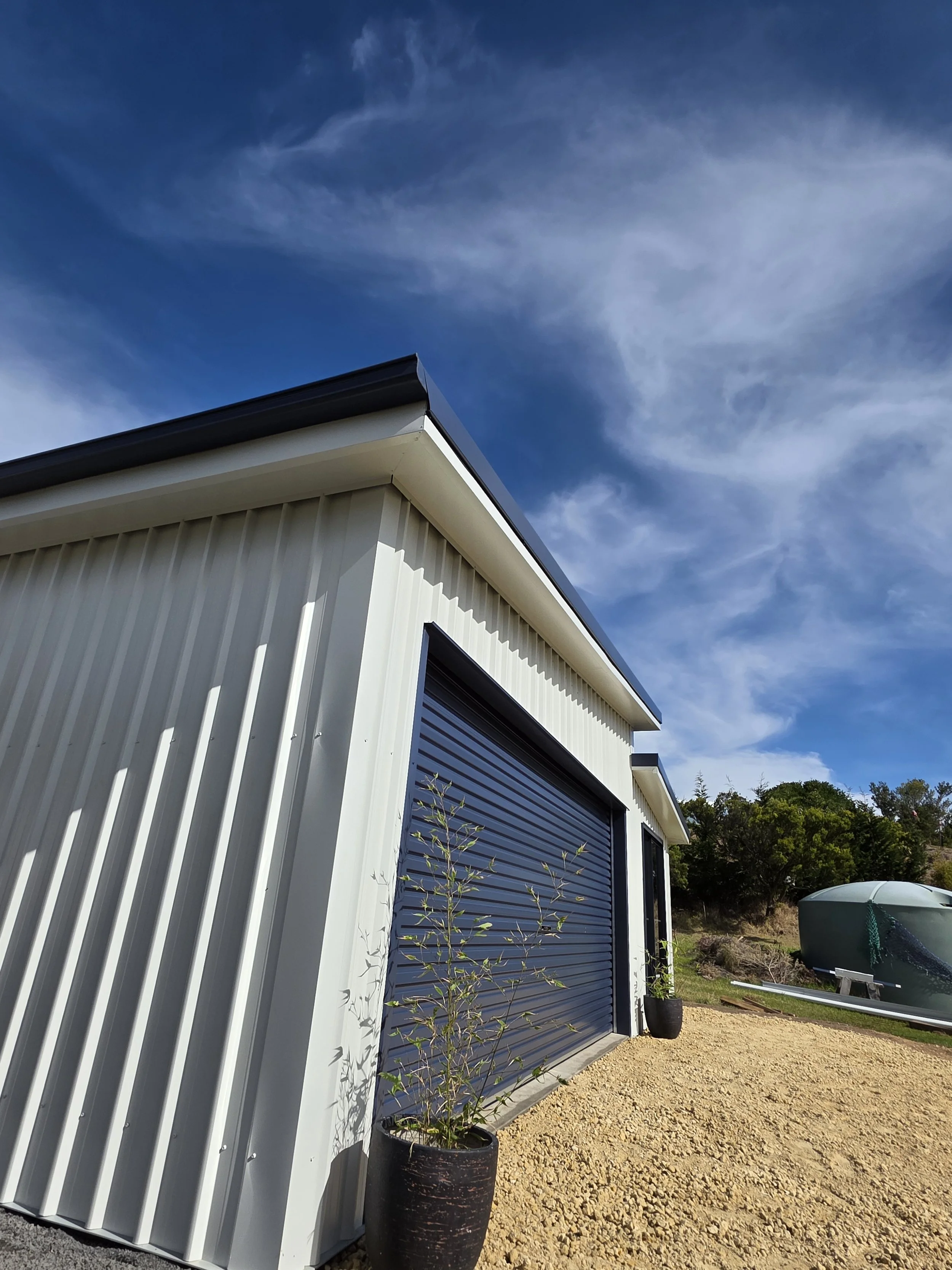 Metal building with a black roll-up door, potted plants outside, and a boat on a trailer nearby under a blue sky with some clouds.