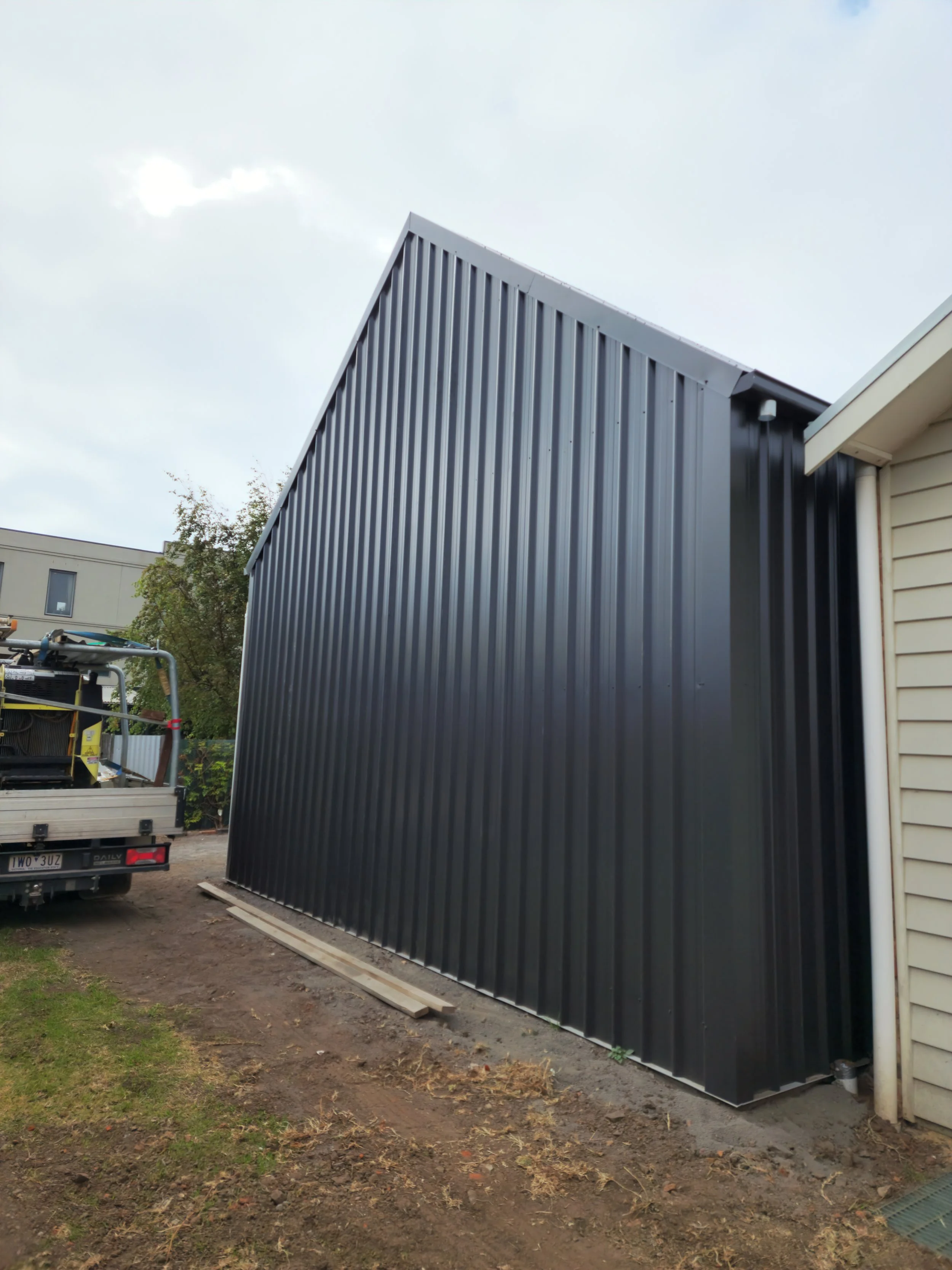 A black metal shed being installed against the side of a house, built by M&C Sheds