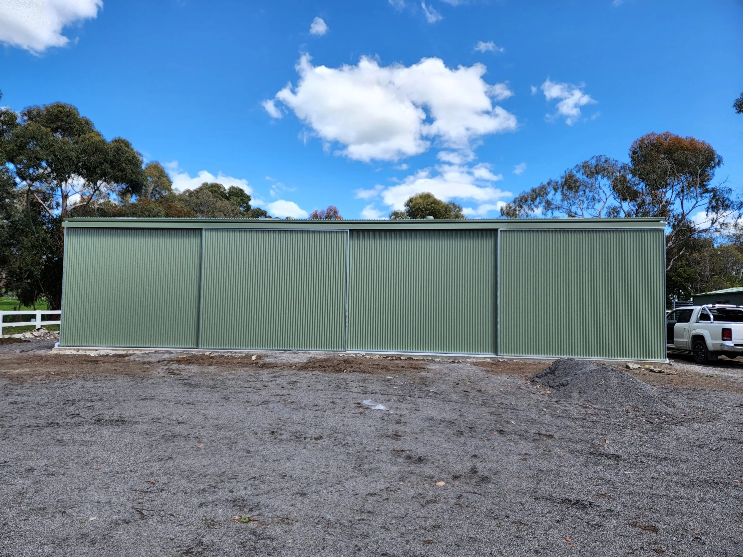 A large green metal shed with sliding doors, surrounded by a gravel area, with trees and a partly cloudy sky in the background.