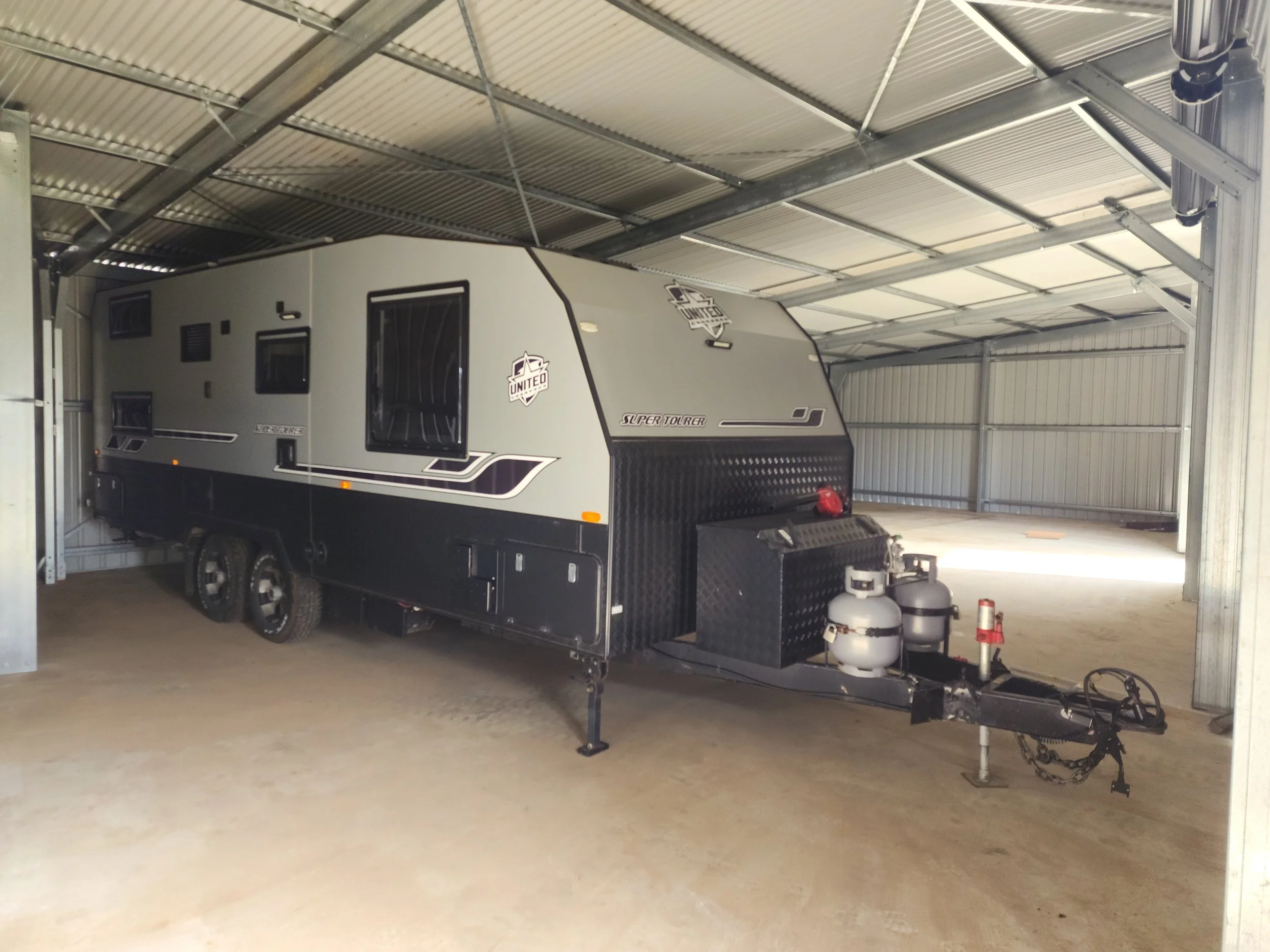 A modern travel trailer parked inside a metal storage shed with a gravel floor, featuring black and gray exterior design, two propane tanks at the front, and black wheels, shed built by M&C Sheds
