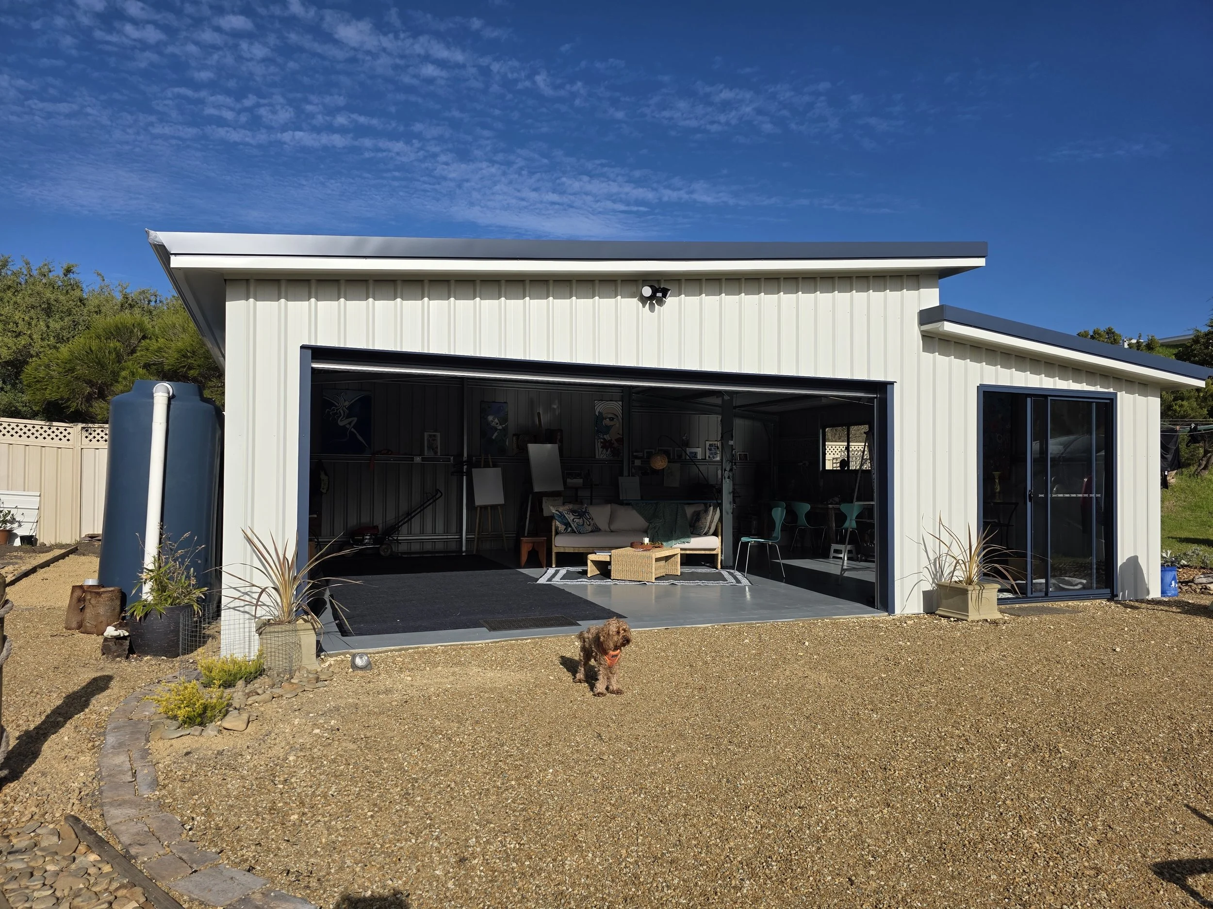 A modern garage with a large open door, containing a seating area with a couch and chairs inside. Outside, a small dog stands on a gravel yard near potted plants, under a clear blue sky with scattered clouds.