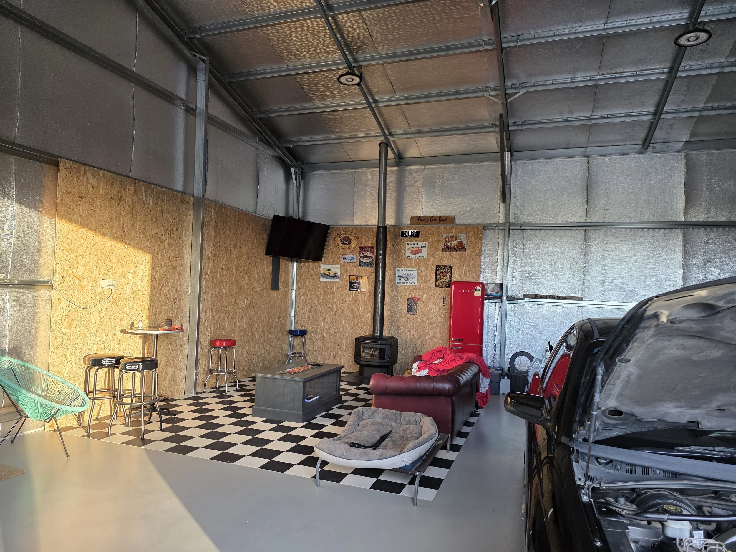 A garage shed with exposed metal walls and ceiling, decorated with vintage car posters, a black flat-screen TV, a red vintage refrigerator, and a wood stove. 