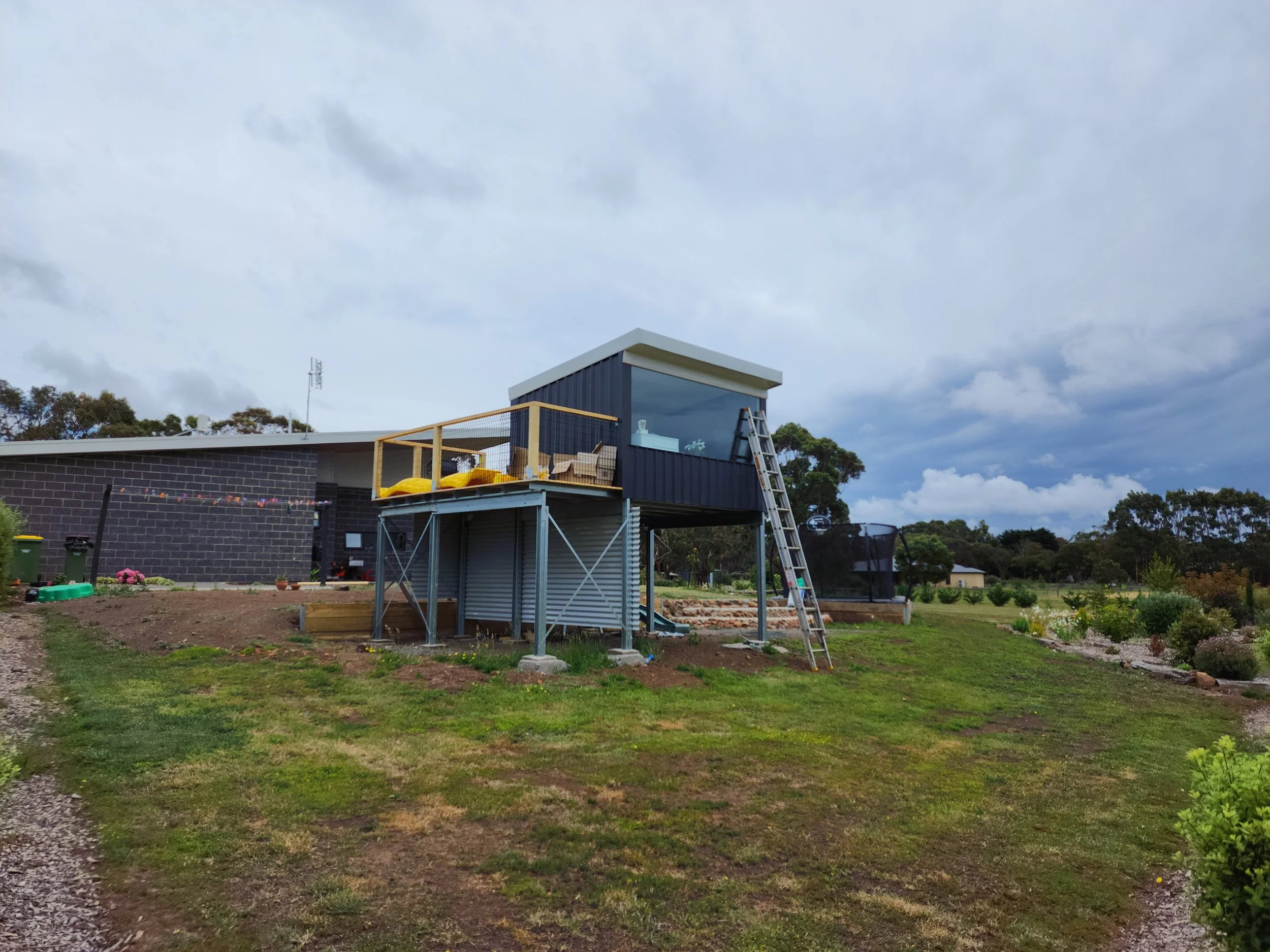 A modern tiny house shed on stilts with a balcony, ladder, and outdoor seating area built by M&C Sheds