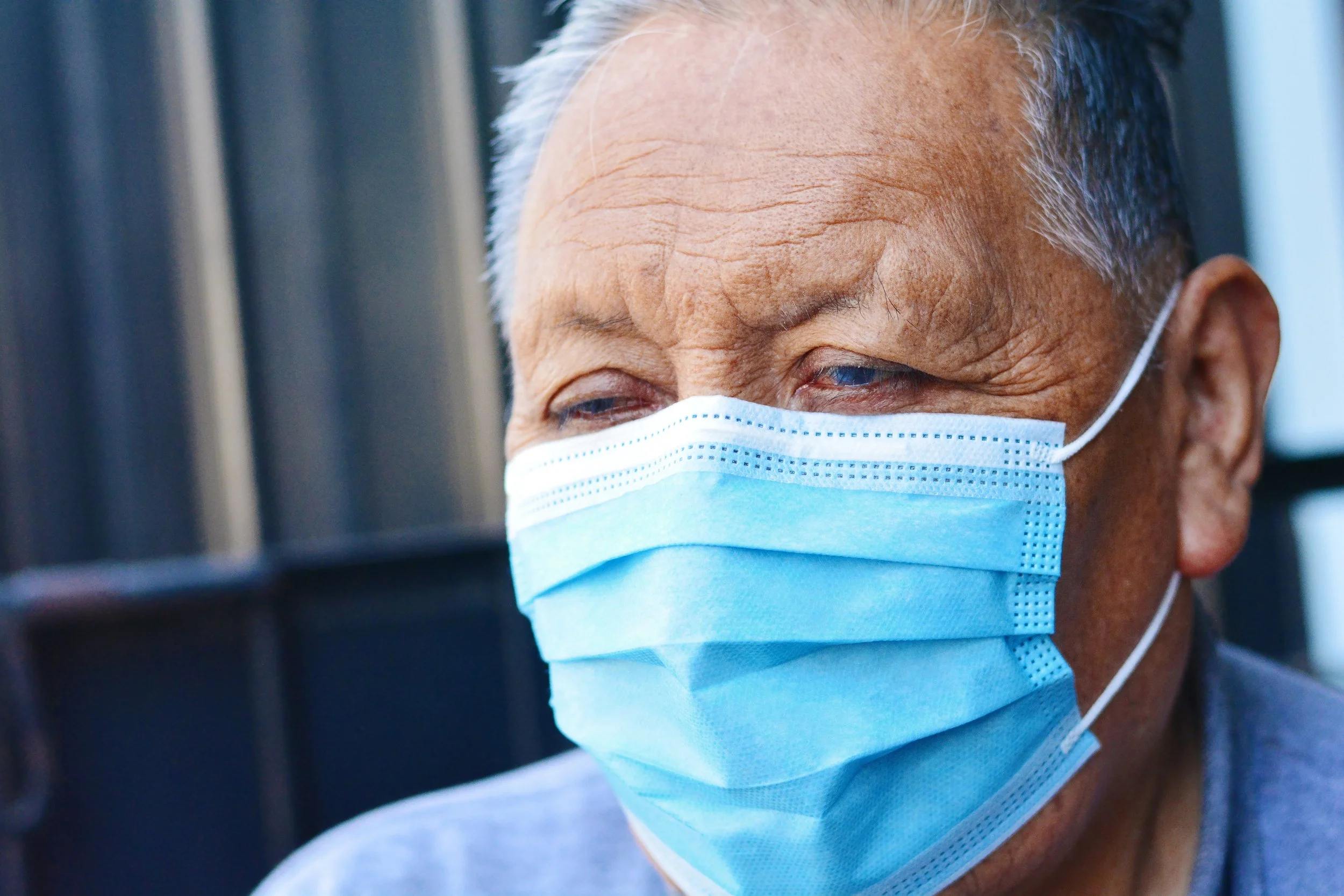 Close-up of an elderly man wearing a blue surgical mask, with gray hair and wearing a blue shirt, in an indoor setting.