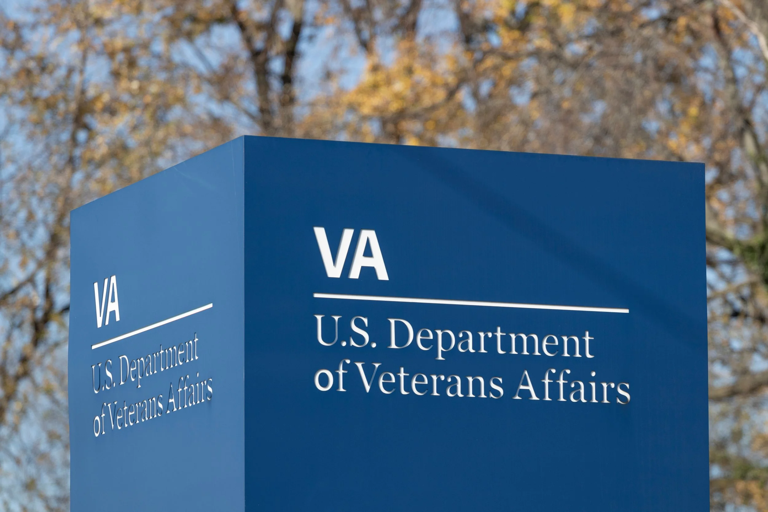 Blue signpost with white text for the U.S. Department of Veterans Affairs, outdoors with trees and blue sky in the background.