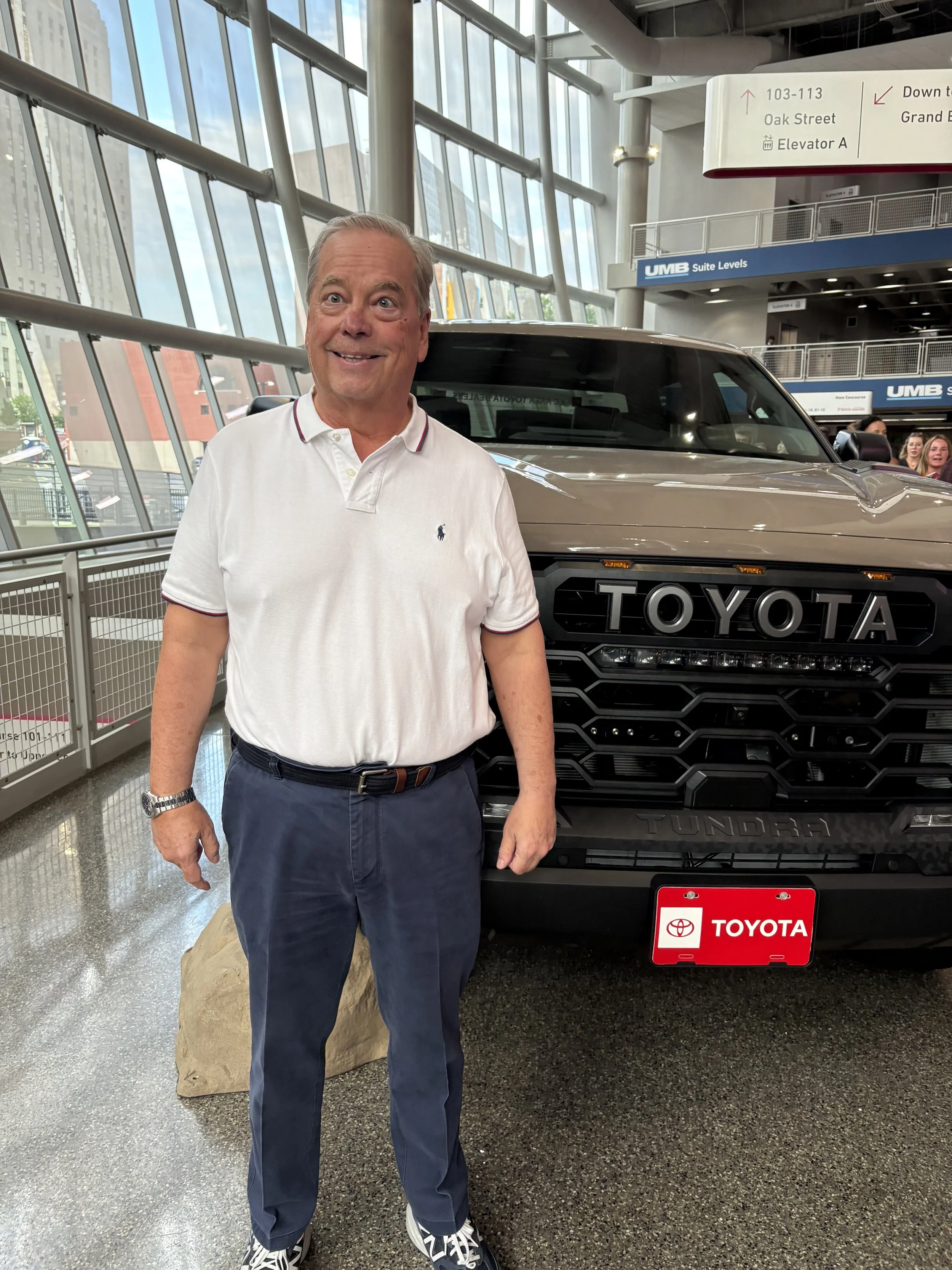 A man standing indoors next to a black Toyota Tundra truck at a car dealership or showroom with large glass windows and a cityscape in the background.