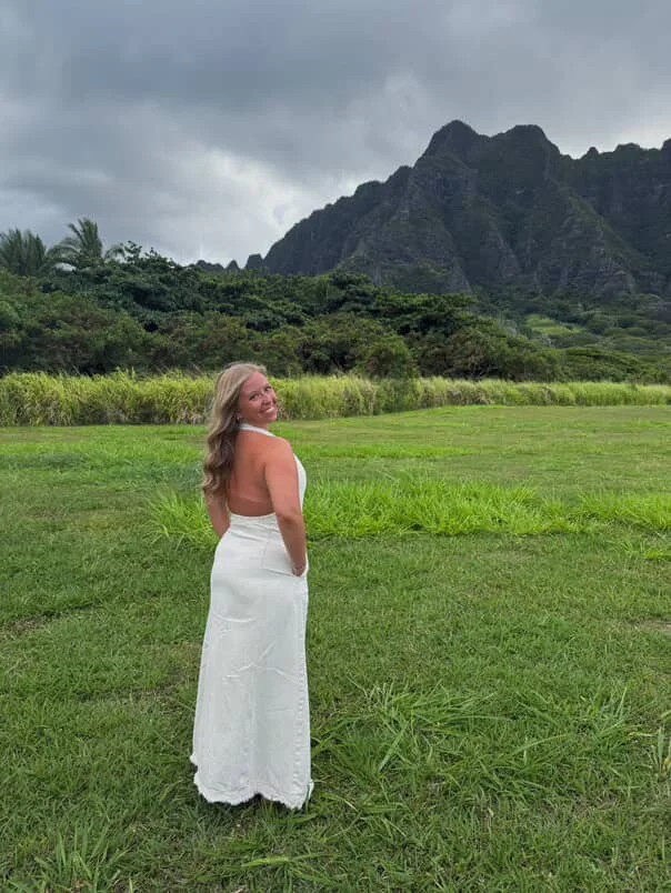 A woman in a white sleeveless dress stands on a lush green field, with mountains and a cloudy sky in the background.