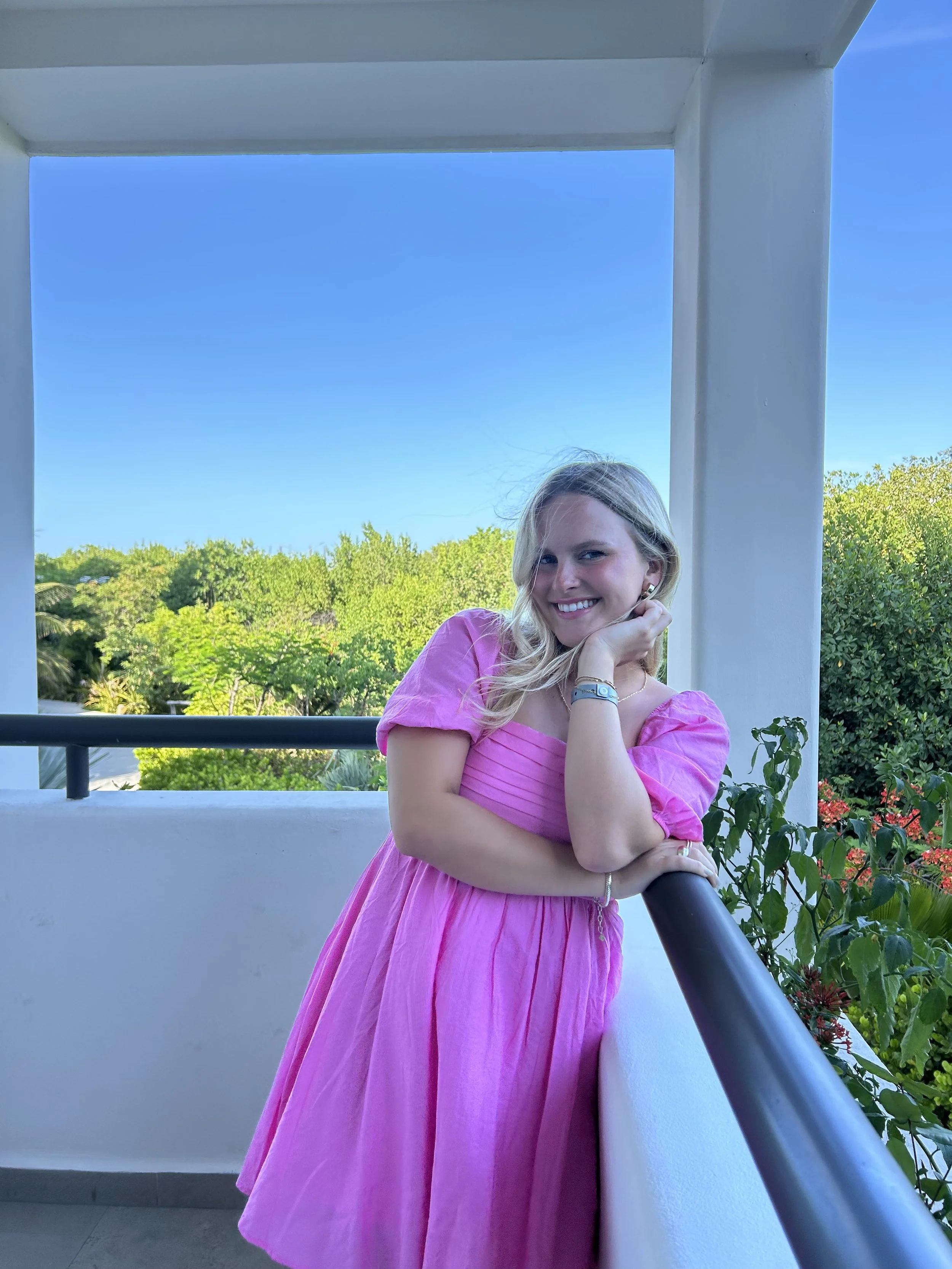 A young woman in a pink dress leaning on a balcony railing, smiling, with green trees and a clear blue sky in the background.