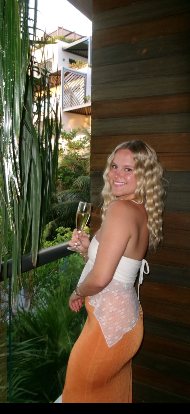 Smiling woman with curly blonde hair holding a glass of champagne, standing on a balcony with lush greenery and modern buildings in the background, wearing a white lace top and orange skirt.