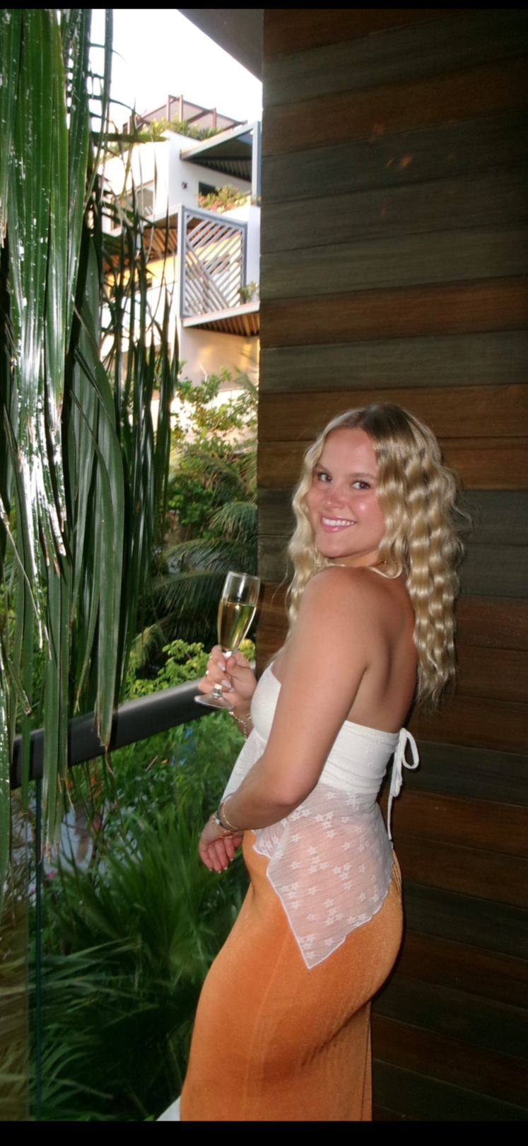 A woman with blonde curly hair smiling and holding a glass of champagne, standing outdoors near a wooden wall and green plants.
