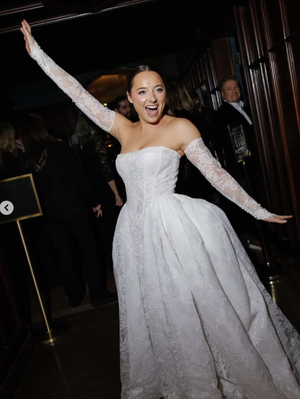A woman in a white wedding dress with long lace sleeves joyfully dancing at her wedding celebration, surrounded by guests in formal attire.