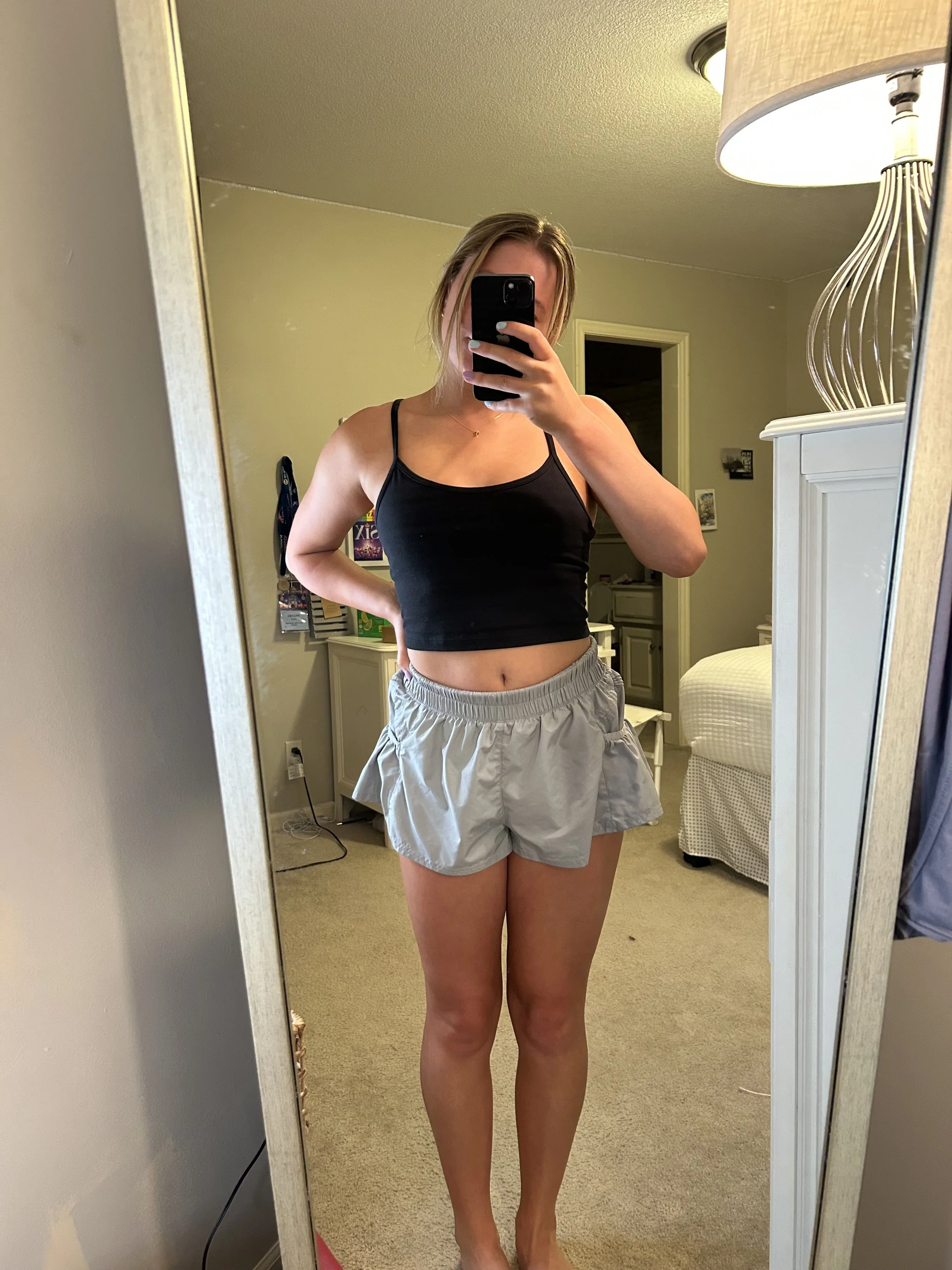 A woman taking a mirror selfie in a bedroom, wearing a black spaghetti strap top and gray shorts.