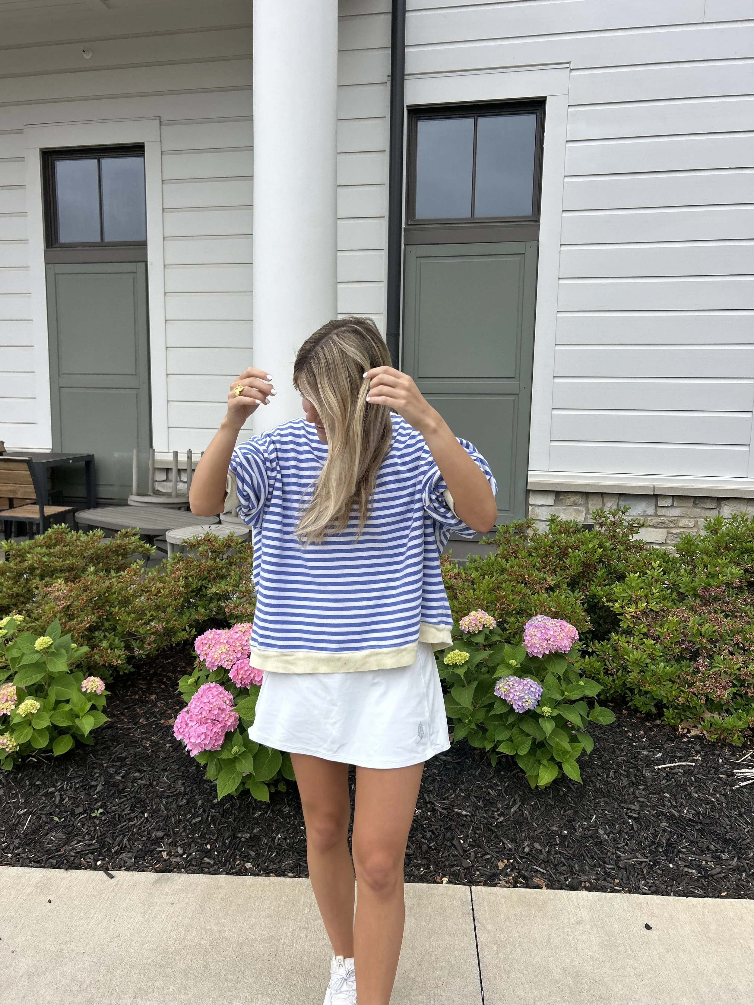 A young woman with long blonde hair standing outdoors in front of bushes with pink and purple hydrangeas. She's wearing a blue and white striped loose top and a white skirt, adjusting her hair with her hands.