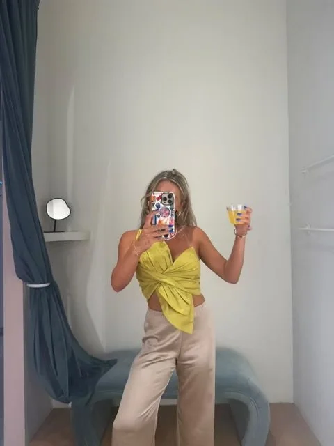 Woman taking a mirror selfie in a room, holding a drink, wearing a yellow top and beige pants.