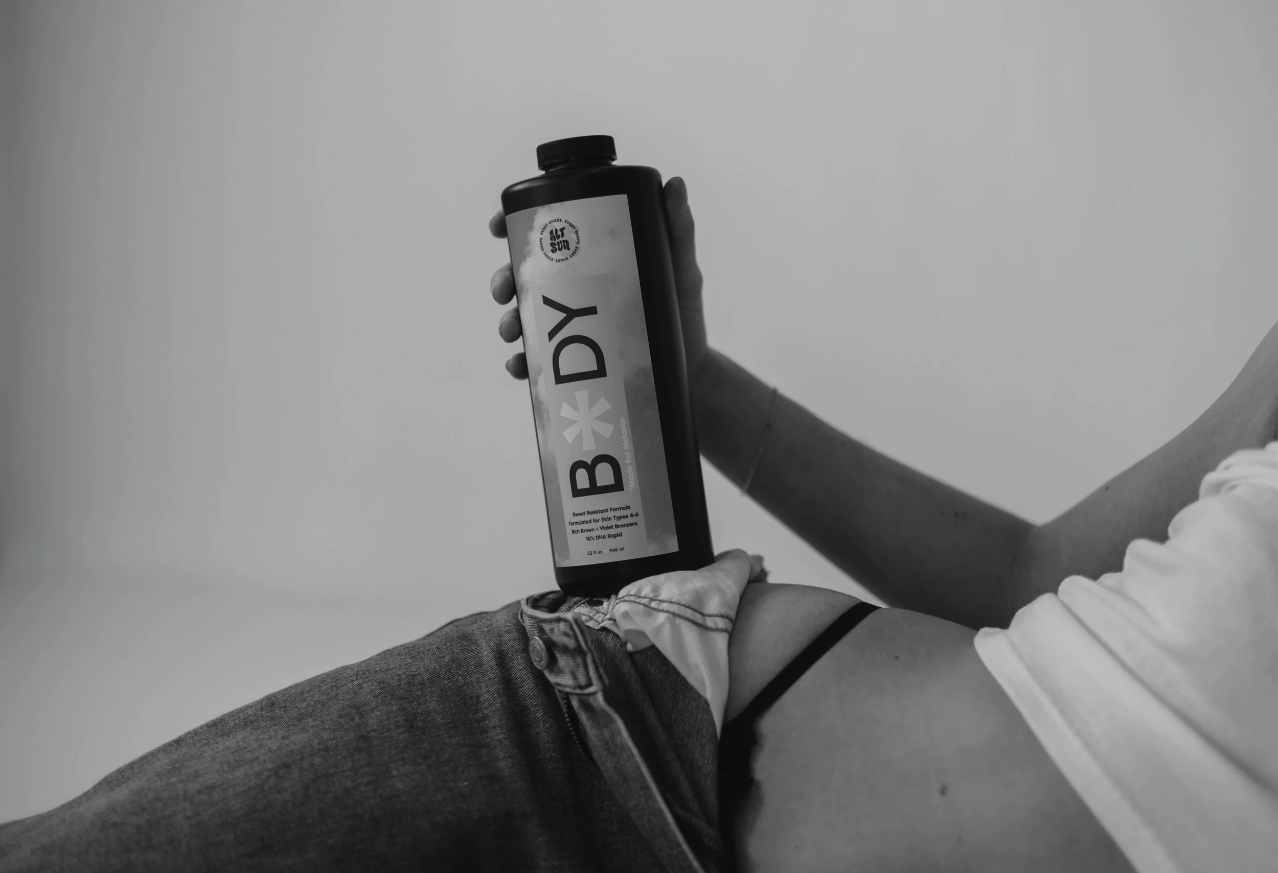 A person holding a black bottle labeled 'BODY' in black and white, with their leg and part of their arm visible, against a plain background.