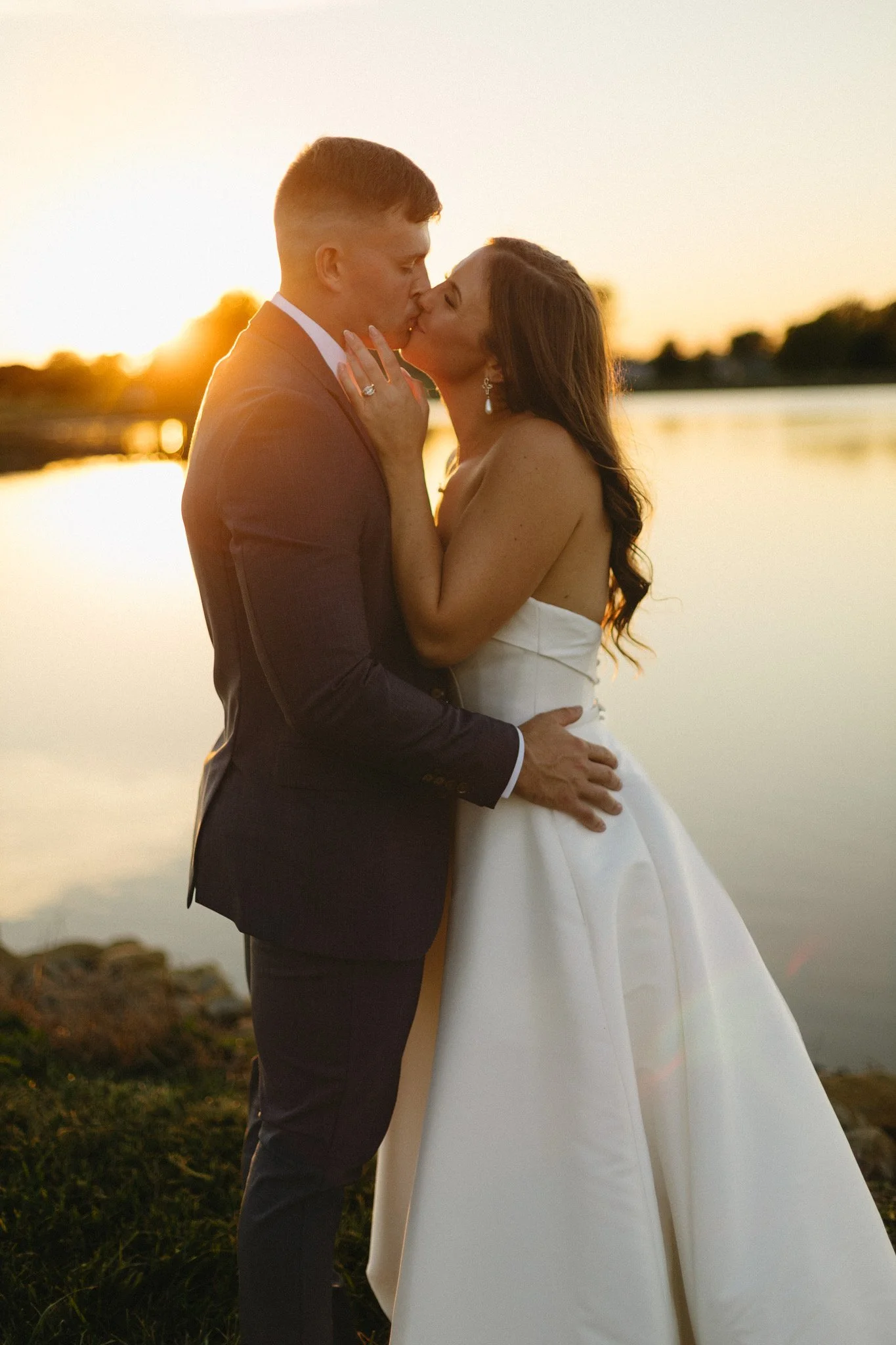 A bride and groom sharing a kiss during sunset by a body of water, with the bride wearing a white gown and the groom in a dark suit.