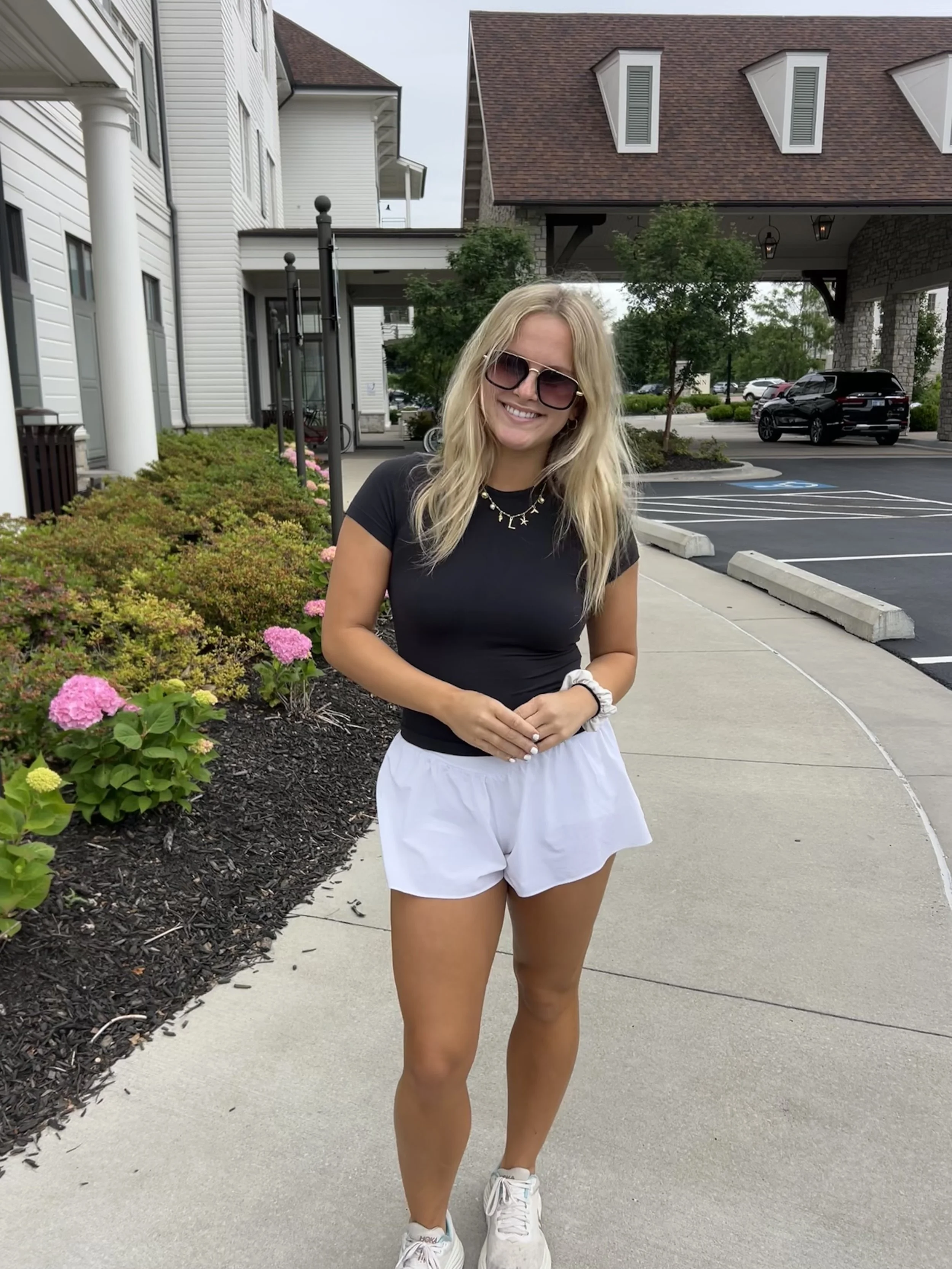 A young woman with blonde hair, wearing sunglasses, a black t-shirt, white shorts, and sneakers, standing on a sidewalk in front of a landscaped area with pink hydrangea flowers, smiling at the camera.