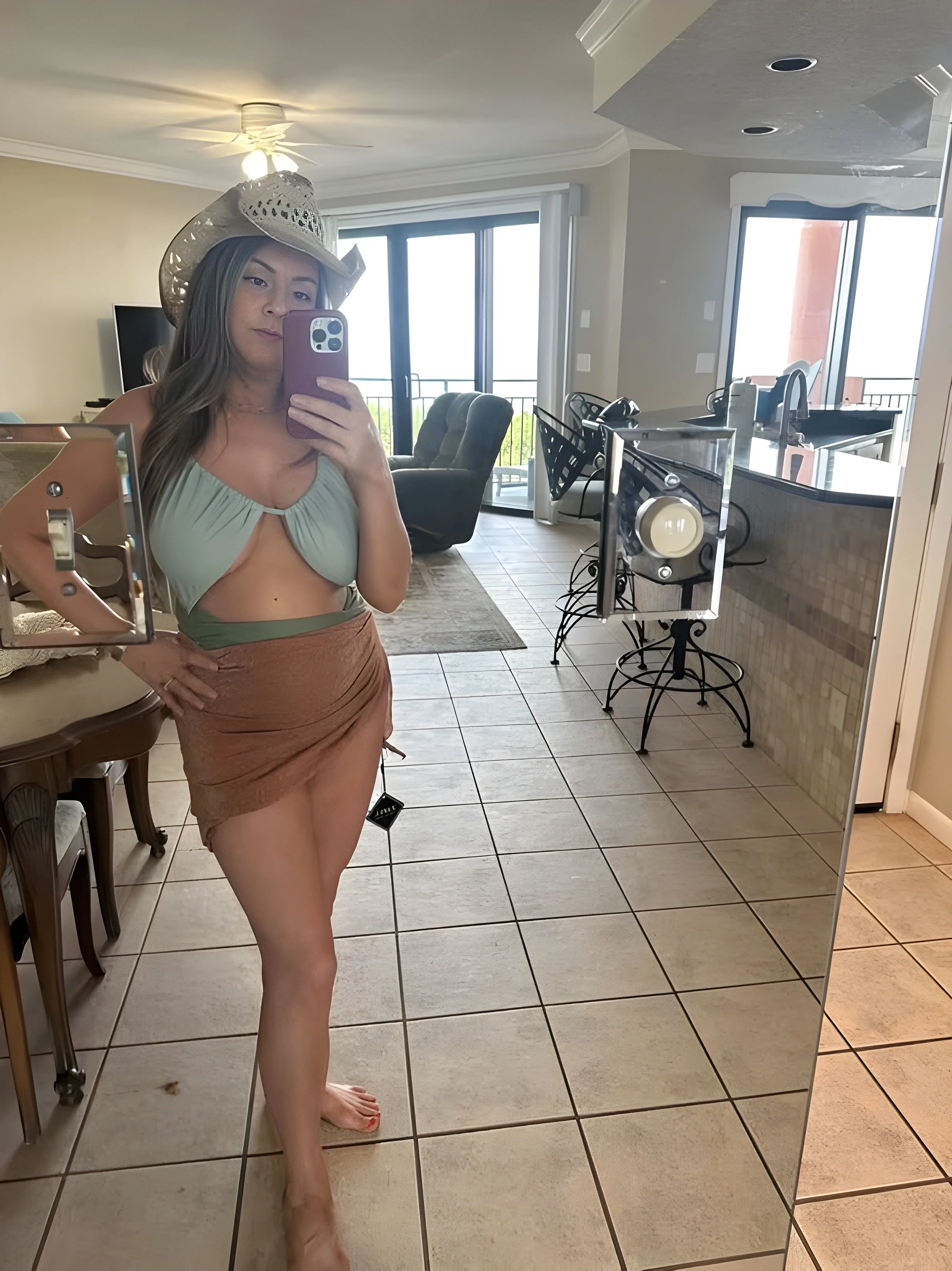 A woman wearing a straw hat, a green top, and a tan skirt taking a mirror selfie in a living room with a balcony view, tiled floor, and modern furniture.