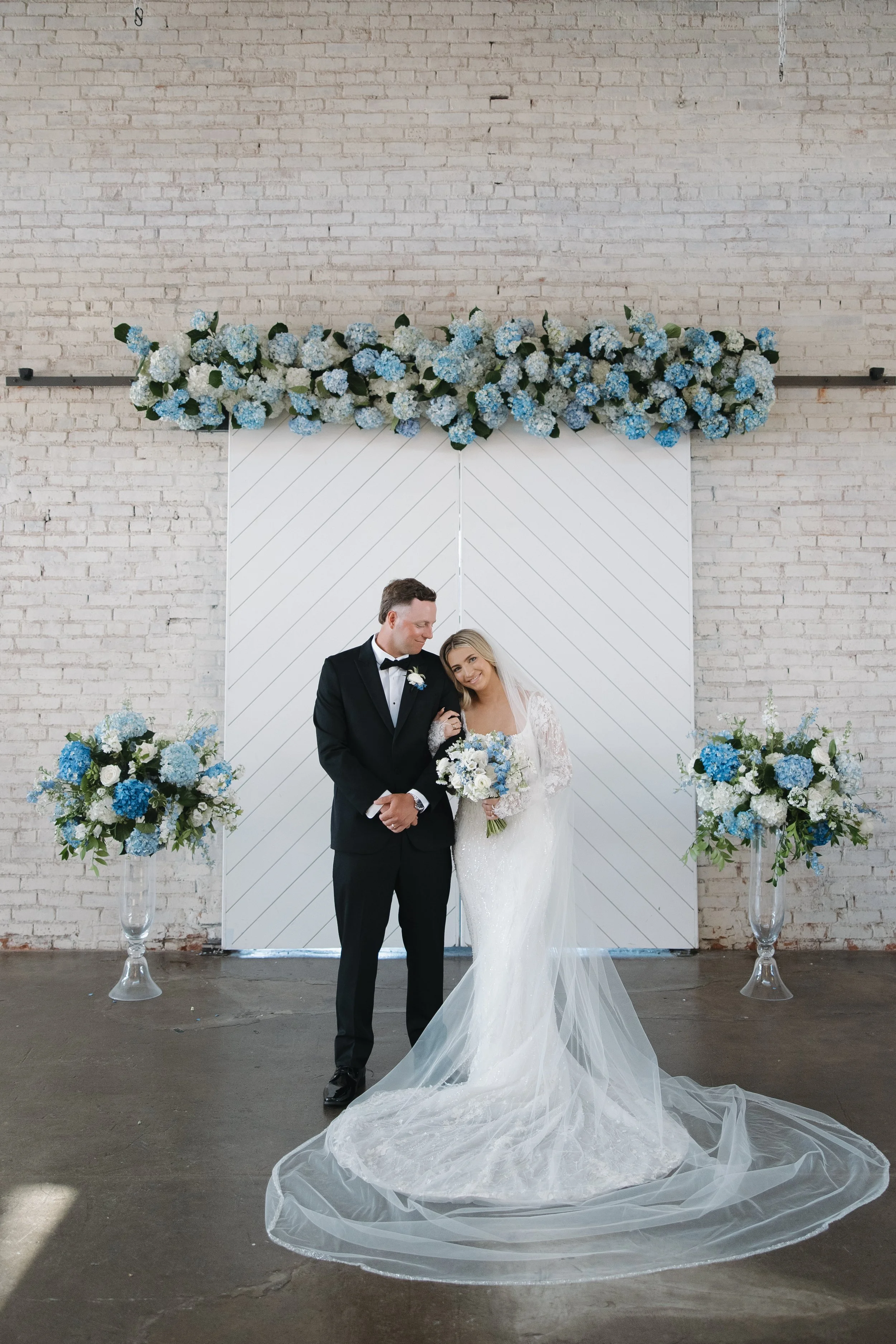 A bride and groom standing together at their wedding, with the bride holding a bouquet, in front of a decorated backdrop with blue and white flowers and a white, striped wall.